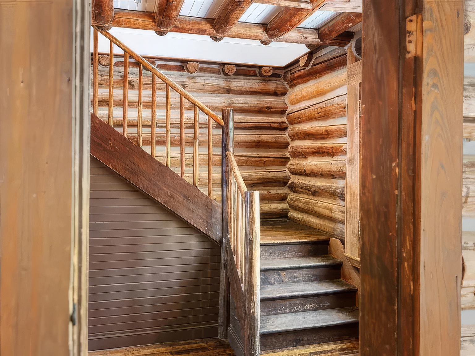 This interior view captures a rustic staircase constructed from natural logs and dark-stained wood, set against walls made of stacked timber logs. The perspective is framed through a doorway, highlighting the textured wood grain and the warm, cabin-like aesthetic of the home. The staircase features a sturdy railing and leads upward, creating a cozy and authentic mountain-lodge atmosphere.