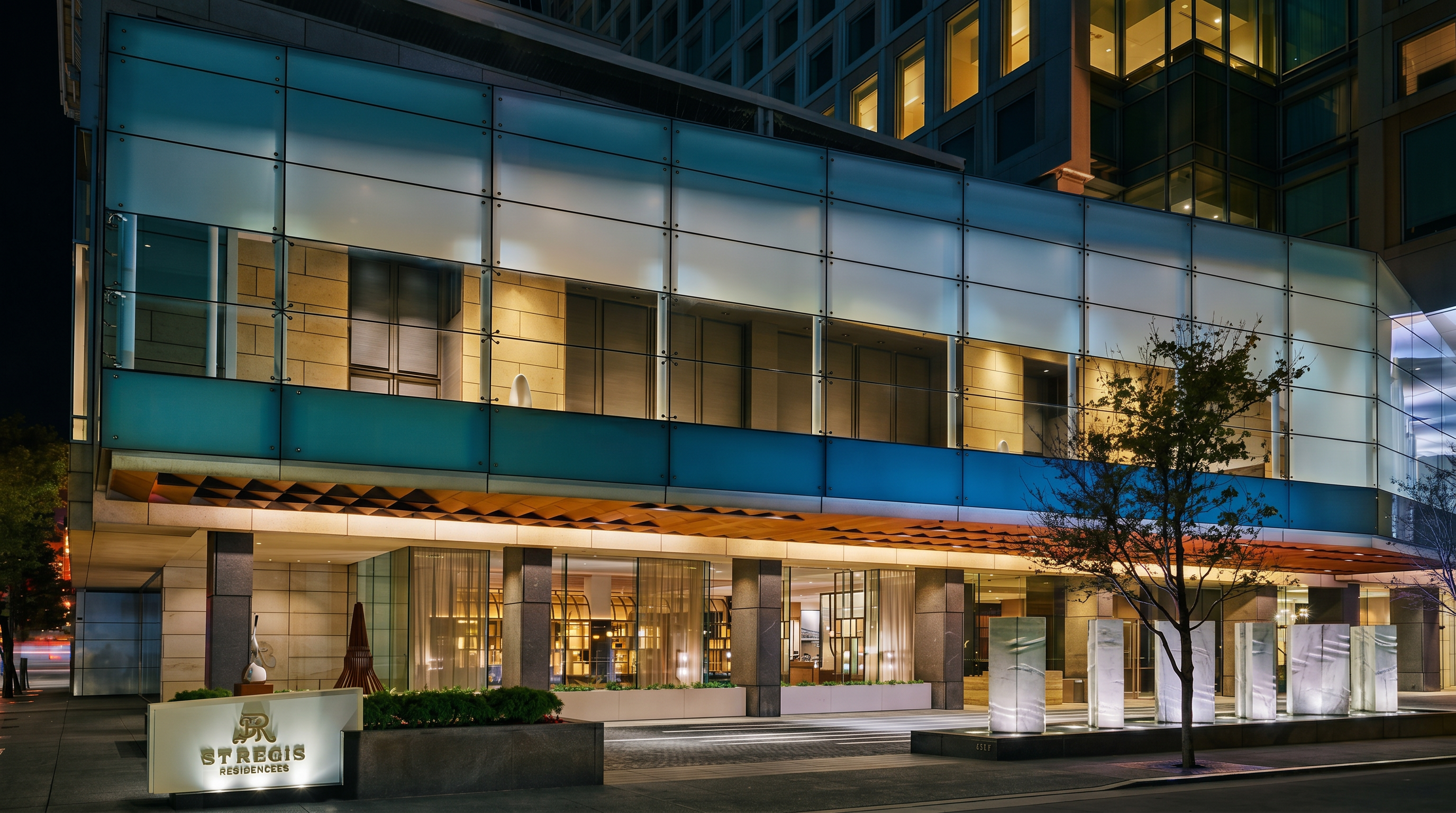 This image captures the sophisticated, modern entrance of the St. Regis San Francisco at night. The building features a prominent glass-paneled facade with warm interior lighting, a stone-clad base, and a series of illuminated sculptural pillars leading to the main entrance. The perspective is a low-angle, eye-level shot that emphasizes the grand scale and contemporary architectural design of the hotel's arrival area.