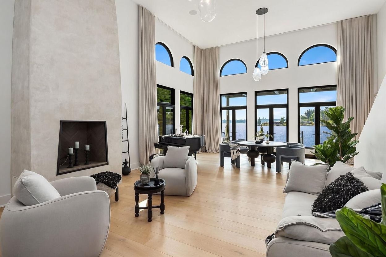 This expansive, light-filled living room features a high-ceiling design with a prominent, tall fireplace surround and large windows that frame a scenic waterfront view. The space is furnished with comfortable, neutral-toned seating, a grand piano, and a circular dining area, creating an elegant and open-concept atmosphere. The light wood flooring and soft, floor-to-ceiling drapery enhance the airy, sophisticated aesthetic of the room.