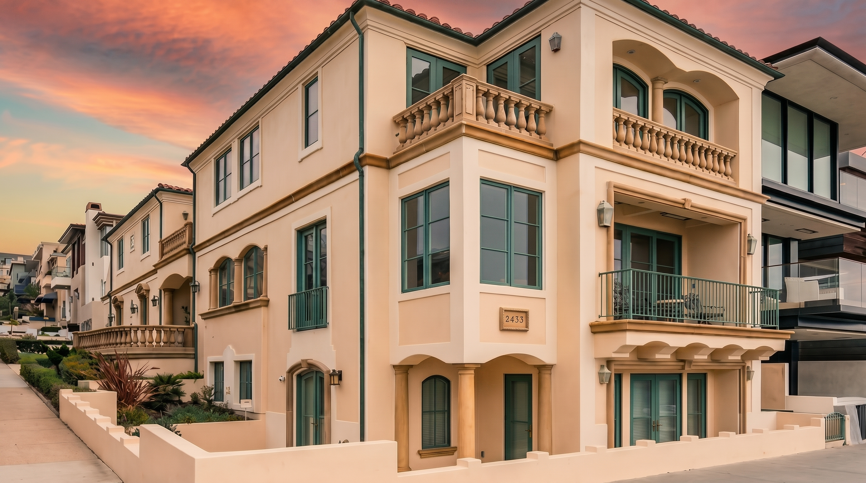 This three-story Mediterranean-style residence features a light-toned stucco facade with elegant architectural details, including arched windows, decorative balustrades, and multiple balconies. The home is situated on a corner lot, showcasing a clean, sophisticated exterior design with a prominent street-level entrance and a sidewalk running along the side. The warm, sunset-hued sky provides a dramatic backdrop, highlighting the building's structured geometry and classic aesthetic.