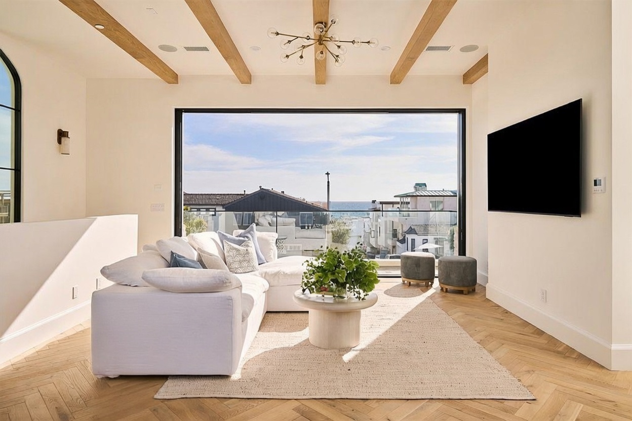 This bright and airy living room features a large, plush white sectional sofa arranged around a circular coffee table on a textured area rug. The space is defined by light wood herringbone flooring and exposed wooden ceiling beams, with a massive wall of glass providing an expansive view of the ocean and coastal neighborhood. A modern chandelier hangs from the ceiling, and a large television is mounted on the wall, creating a sophisticated and relaxing atmosphere.