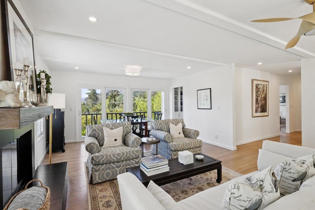 This bright and airy living room features light wood flooring and a neutral color palette, centered around a dark wood coffee table and two patterned armchairs. A fireplace with a dark mantel serves as a focal point on the left, while large glass sliding doors provide ample natural light and a view of the outdoor balcony. The space flows seamlessly into an adjacent dining area, creating an open-concept layout ideal for entertaining.