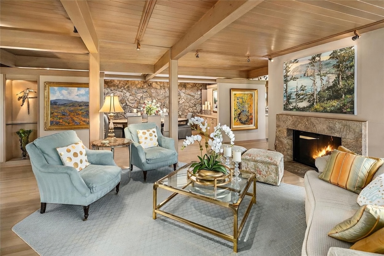 This elegant living room features a warm, wood-paneled ceiling with exposed beams and a stone-clad fireplace surround that serves as a focal point. The space is furnished with light blue armchairs, a curved sofa, and a central glass-topped coffee table, all arranged on a soft-toned area rug. A dining area with a stone wall is visible in the background, creating an open-concept feel that emphasizes the home's sophisticated, mid-century inspired aesthetic.