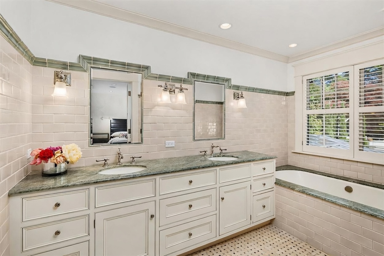 This primary bathroom features a dual-sink vanity with a green stone countertop and white cabinetry, complemented by a tiled backsplash that extends along the wall. A deep soaking tub is positioned beneath a window with shutters, and the room is finished with a patterned tile floor. The perspective is a wide-angle shot capturing the entire vanity area and the adjacent tub, creating a bright and classic aesthetic.