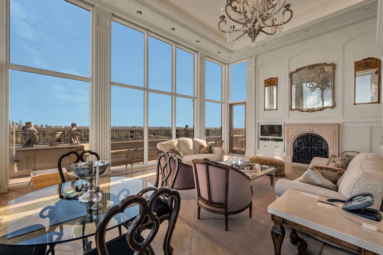 This elegant living and dining area features expansive floor-to-ceiling windows that provide a panoramic view of the city skyline and terrace. The room is furnished with sophisticated, traditional-style seating, a glass-topped dining table, and a grand crystal chandelier hanging from the high ceiling. A stone fireplace surround serves as a focal point on the wall, complemented by ornate mirrors and classic architectural molding.