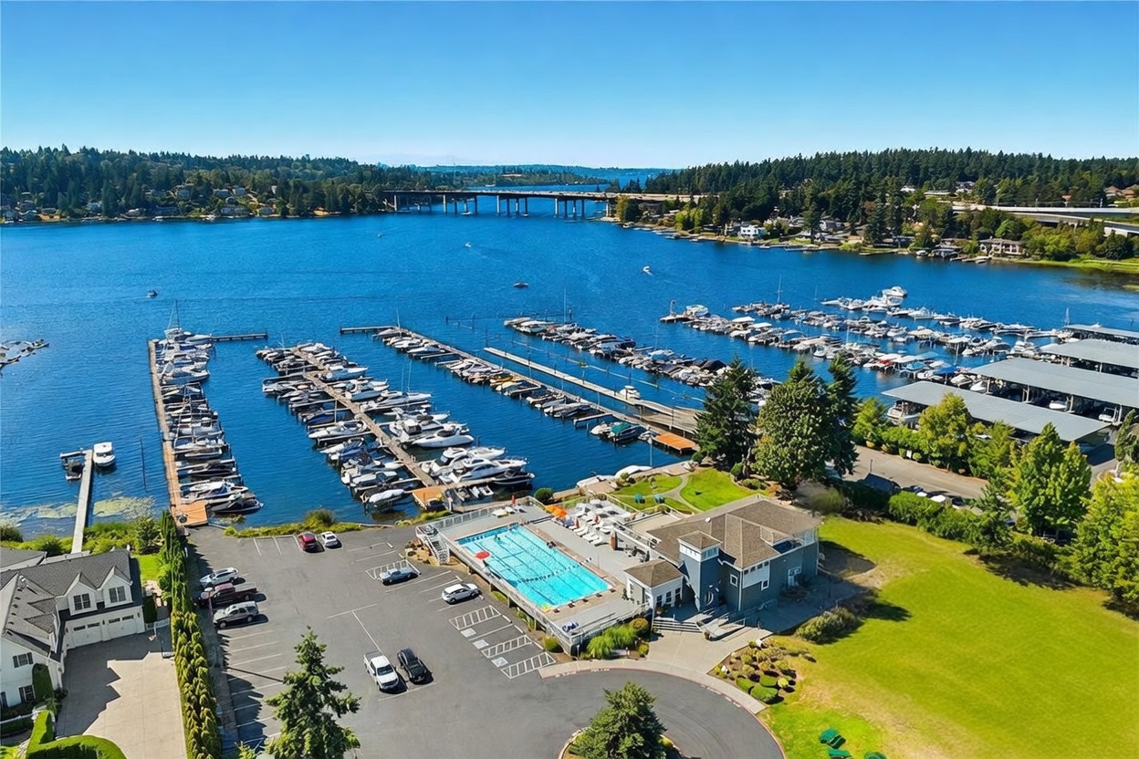 This high-angle aerial view captures a waterfront community featuring a large marina filled with boats, a swimming pool, and tennis courts. The property is situated along a scenic lake with a bridge visible in the background, surrounded by lush greenery and residential homes. The perspective highlights the expansive recreational amenities and the prime location of the community.