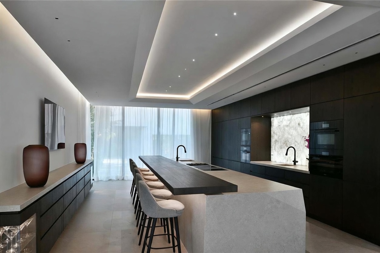 This sleek, modern kitchen features a large central island with a dark wood breakfast bar and a light-toned stone base, complemented by a row of matching bar stools. The space is defined by dark, minimalist cabinetry, integrated appliances, and a striking backlit stone backsplash. A recessed ceiling with integrated lighting adds a sophisticated, cinematic glow to the room, which opens up to a bright area with floor-to-ceiling curtains.