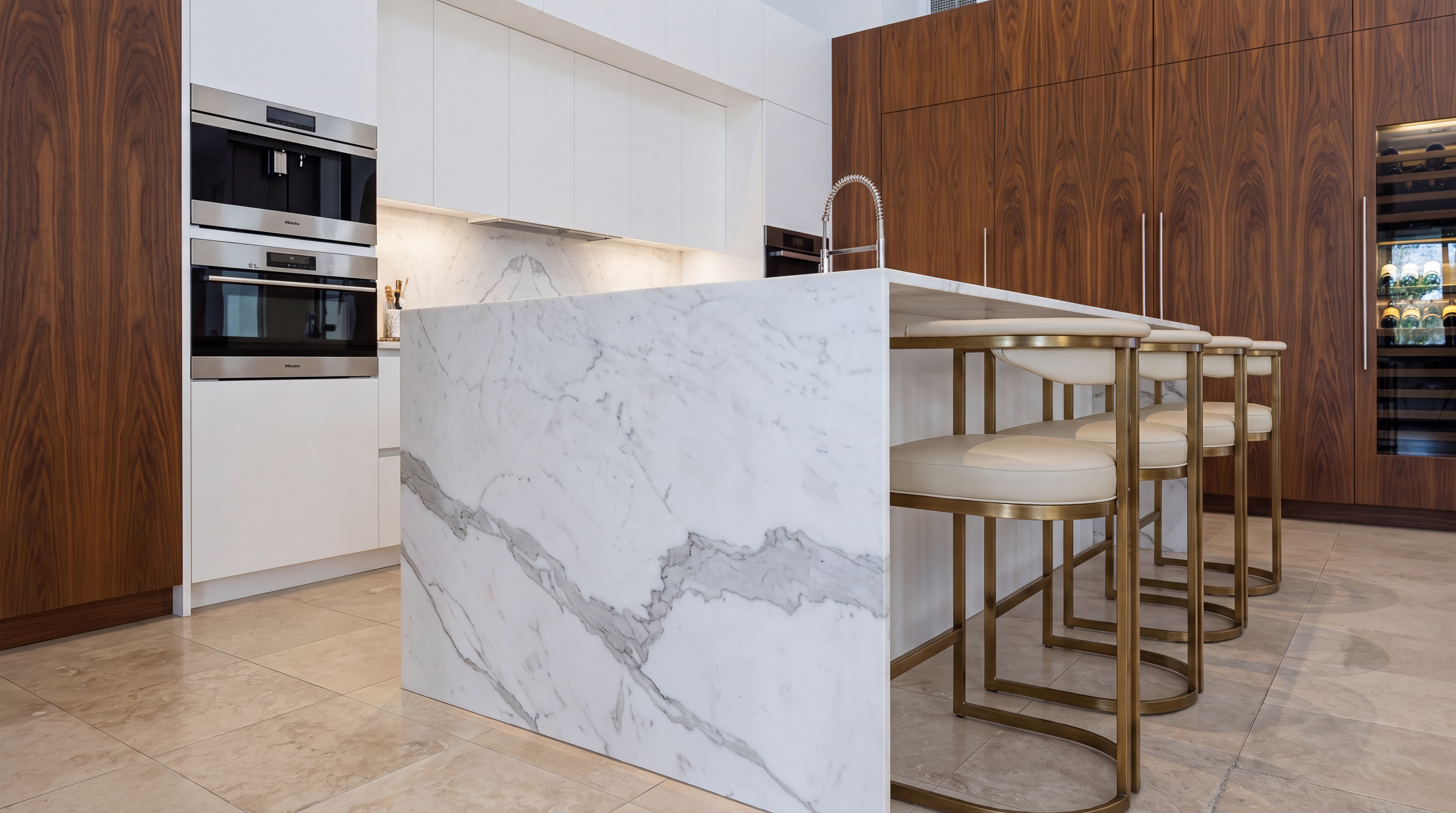 This stunning contemporary kitchen features a seamless blend of sleek white flat-panel cabinetry and warm walnut wood paneling. The centerpiece is a substantial waterfall-edge marble island equipped with a integrated sink, complemented by stylish brass-framed bar stools. These high-end finishes, combined with premium Miele appliances and a built-in wine cooler, create an atmosphere of refined luxury and modern sophistication.