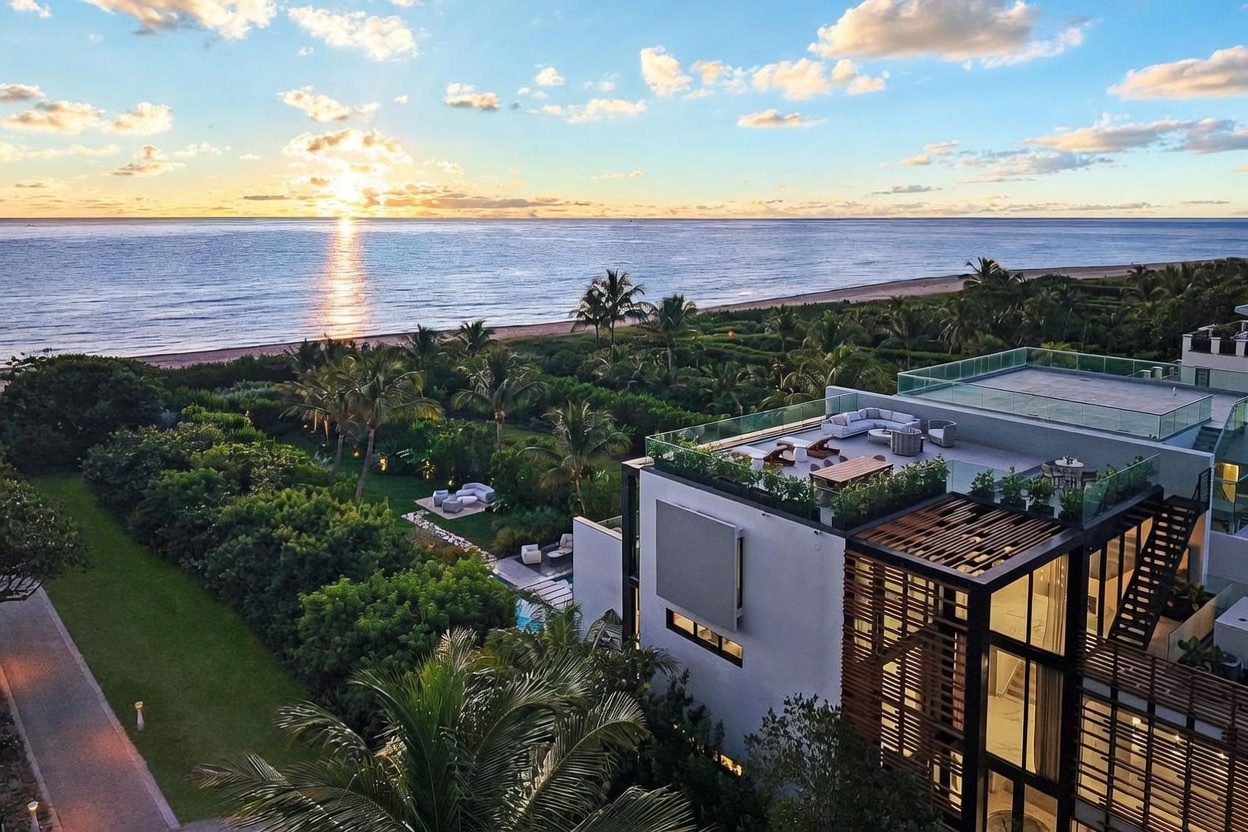 This high-angle aerial shot captures a luxurious modern residence situated directly along a pristine coastline at sunset. The property features a spacious rooftop terrace with lounge seating and dining areas, overlooking lush tropical landscaping and a private pool area below. The expansive ocean view and the warm, golden light of the setting sun create an atmosphere of sophisticated coastal elegance.