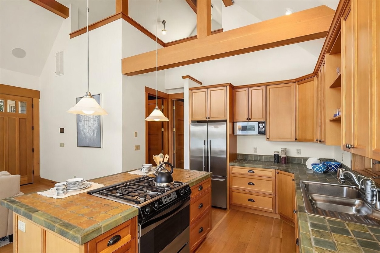 This warm and inviting kitchen features natural wood cabinetry, tiled countertops, and a central island equipped with a gas cooktop. The space is highlighted by high vaulted ceilings with exposed wooden beams and two pendant lights hanging over the island, creating an open and airy atmosphere. The perspective is a wide-angle shot from the kitchen entrance, showcasing the functional layout and the seamless integration of the cooking and preparation areas.