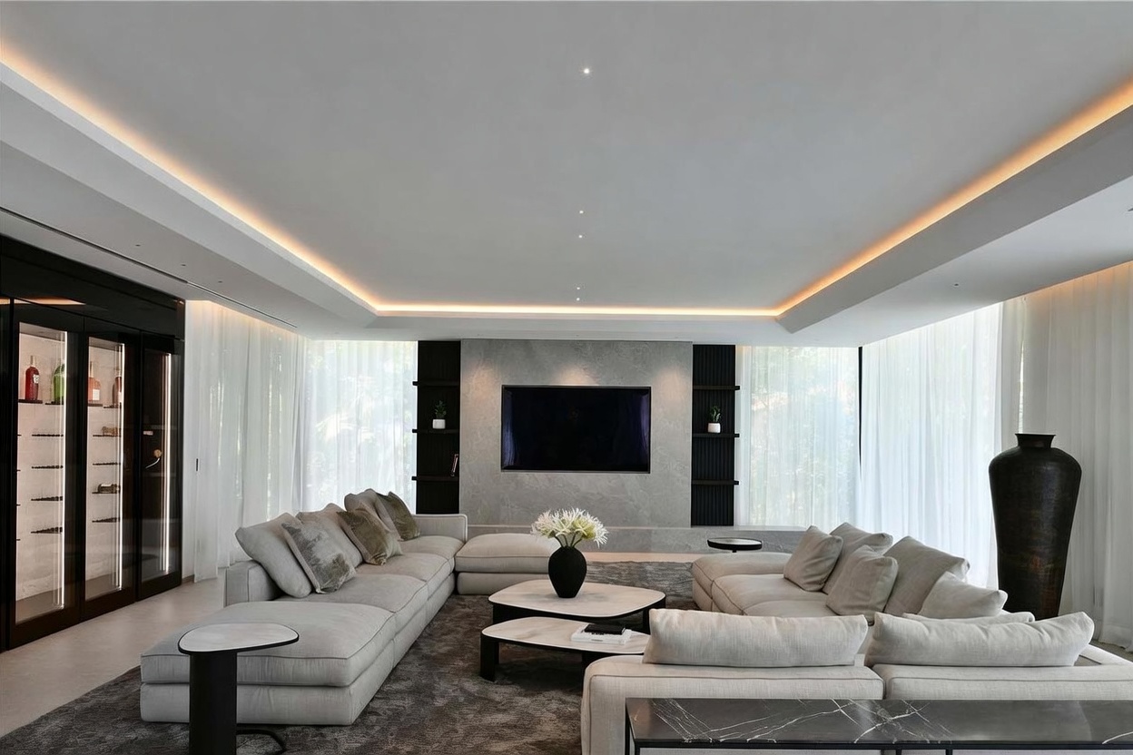 This sophisticated living room features a large, neutral-toned sectional sofa arranged around a pair of tiered coffee tables on a plush area rug. A central wall showcases a mounted television set against a textured stone-clad chimney breast, flanked by dark, built-in shelving units. The room is illuminated by recessed lighting and a warm, perimeter cove light, creating an inviting atmosphere, while sheer curtains frame the large windows on either side.