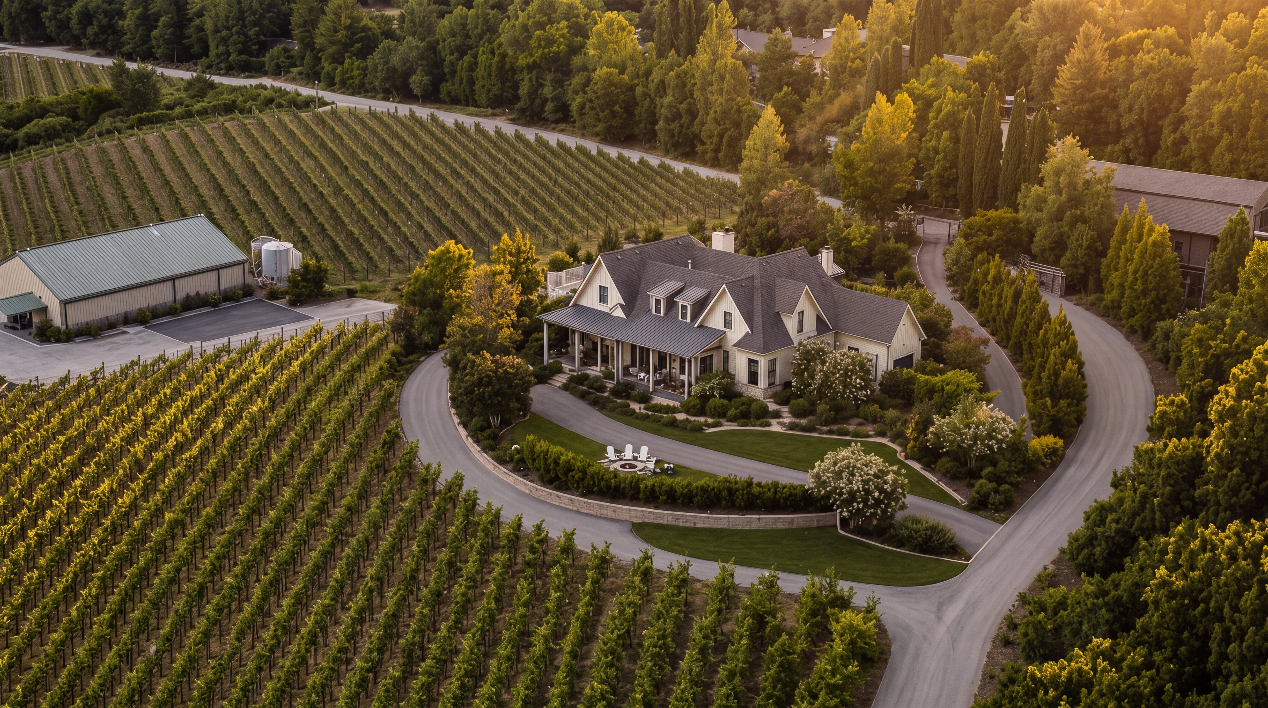 This stunning aerial view captures an expansive estate surrounded by meticulously maintained vineyards and lush greenery. The centerpiece is a classic farmhouse-style residence with a dark metal roof, large porch, and a private circular driveway featuring a fire pit area. The property includes a large secondary building and mature landscaping, presenting a serene, luxurious country living environment.