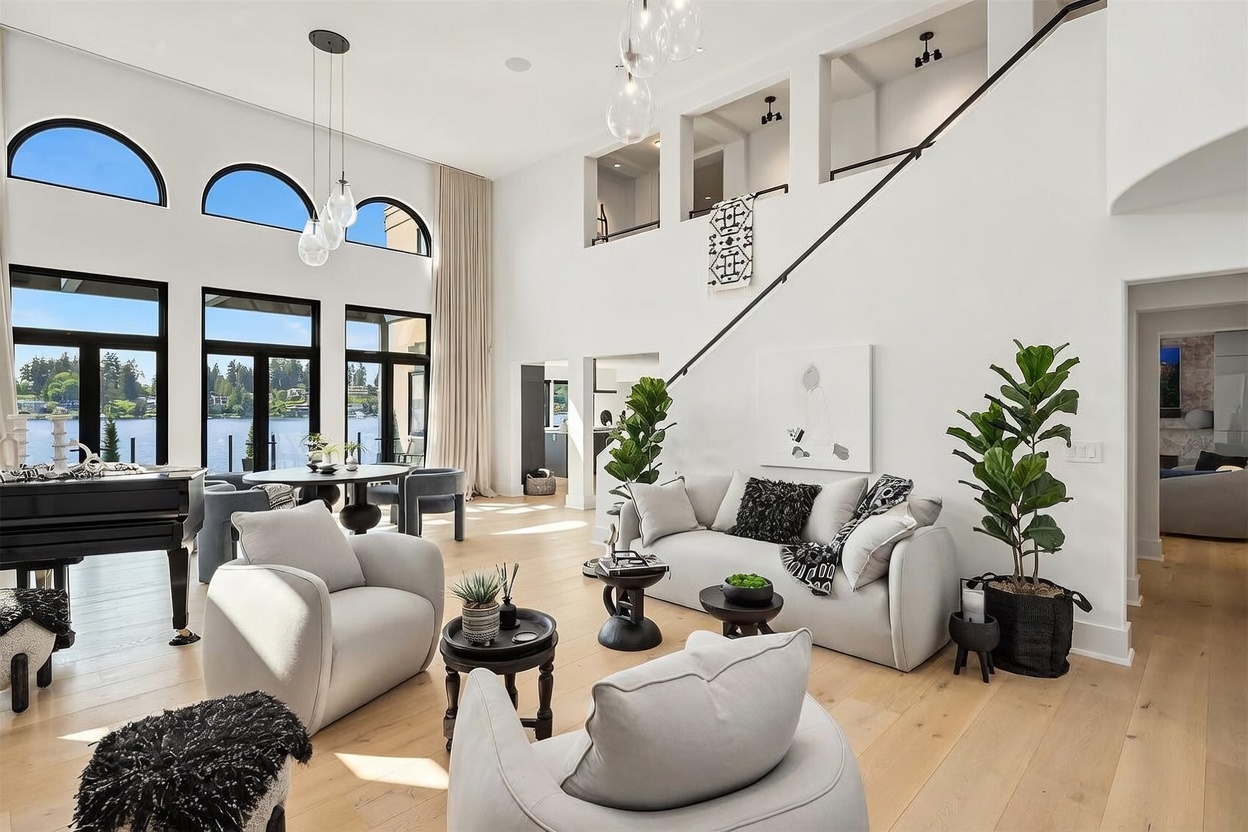This expansive, light-filled living room features a double-height ceiling and a wall of large windows that provide a scenic view of the water. The space is furnished with a contemporary sofa, two plush armchairs, and a grand piano, all arranged on light wood flooring. A staircase with a sleek railing ascends along the wall, while modern pendant lights and potted plants add a sophisticated, airy touch to the open-concept design.