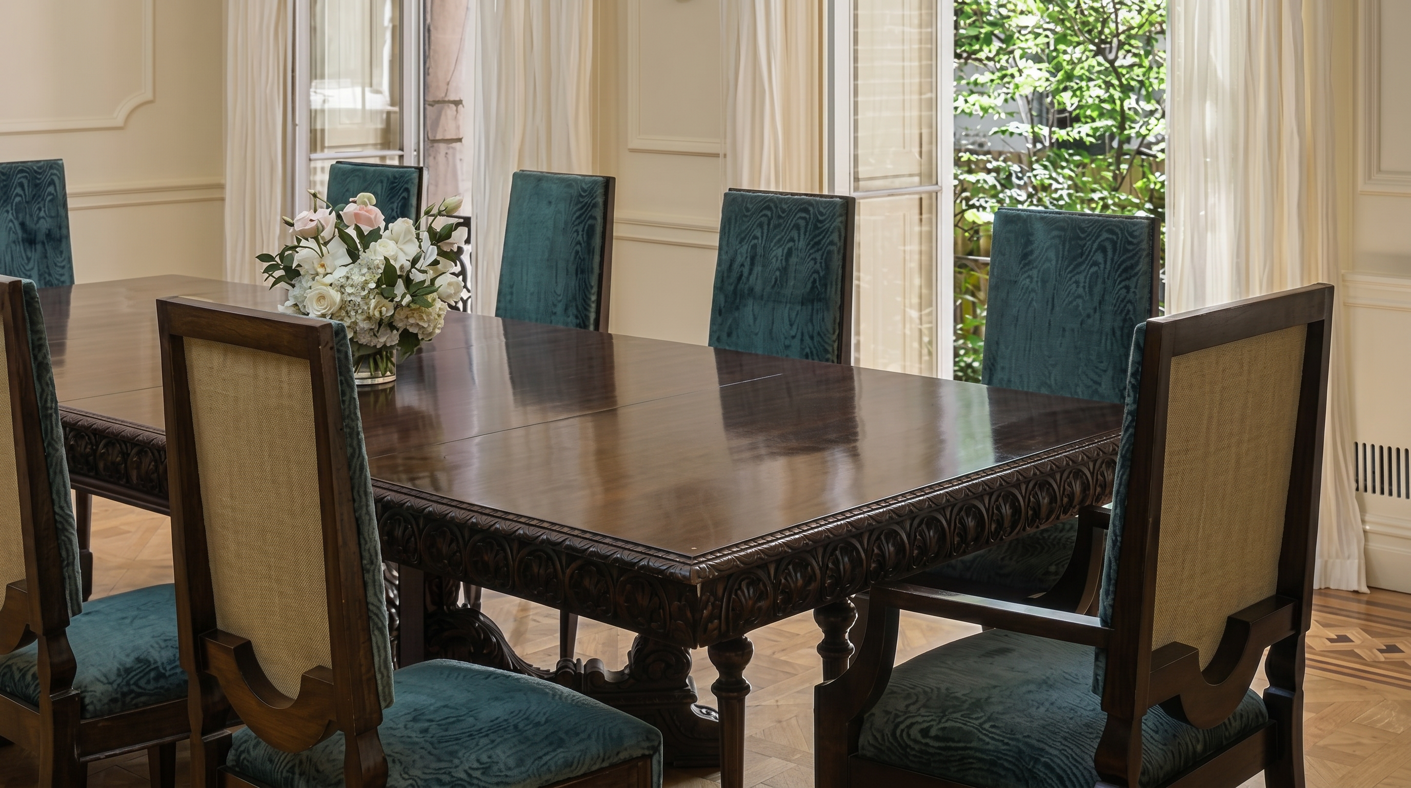 This elegant dining room features a large, dark wood table with intricately carved detailing, surrounded by sophisticated chairs upholstered in patterned teal fabric. The setting is brightened by natural light streaming through large windows draped in simple white curtains, while a decorative floral centerpiece adds a touch of refinement. The classic molding and polished floors underscore the formal and luxurious aesthetic of the space.