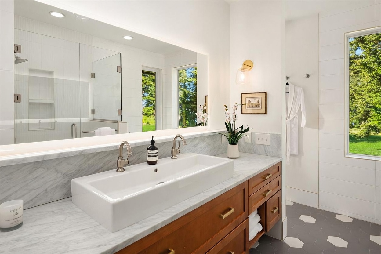 This bright and modern bathroom features a long, rectangular dual-faucet sink set into a stone countertop with a warm-toned wood vanity. A large, wide mirror spans the wall above the vanity, reflecting the shower area and a window that brings in natural light. The space is finished with a geometric patterned floor and clean white wall tiles, creating a polished and serene atmosphere.
