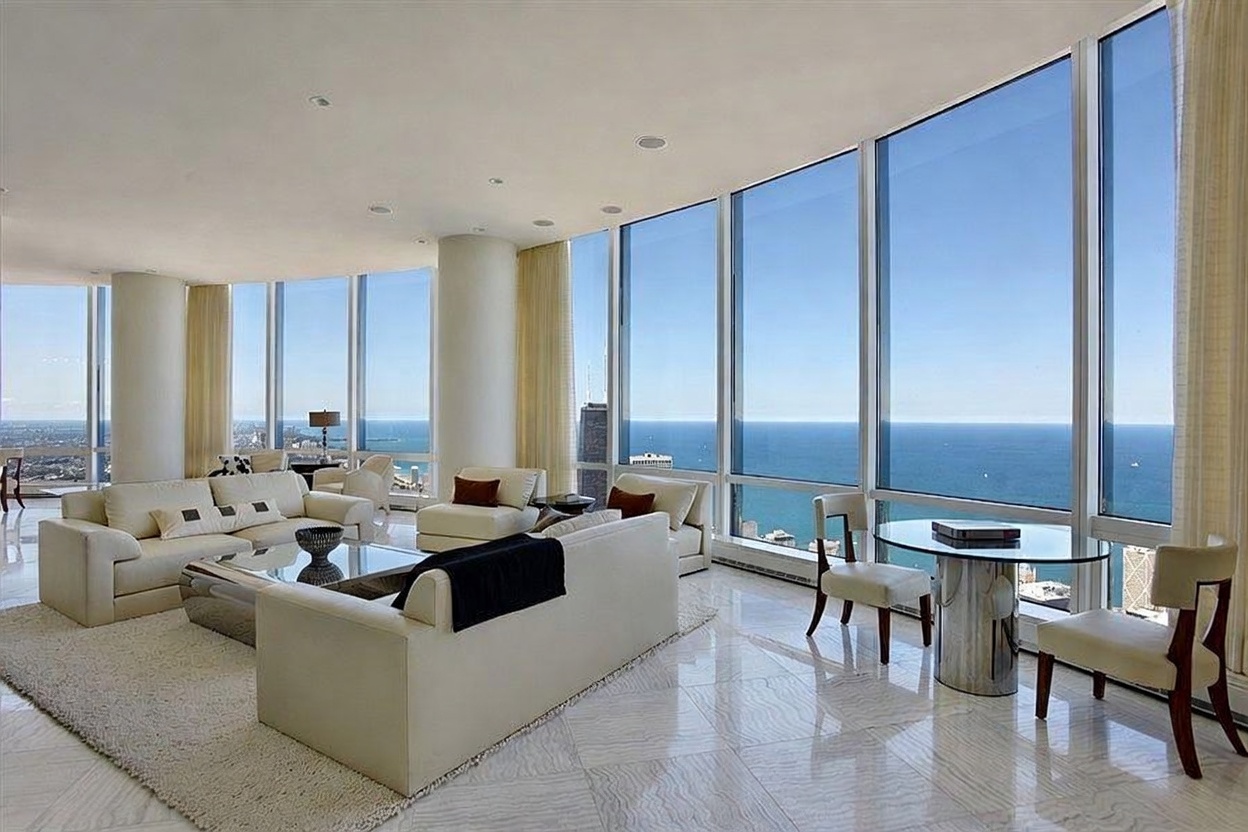 This expansive, high-rise living room features a bright, open-concept layout with polished stone flooring and a neutral color palette. The space is defined by massive floor-to-ceiling windows that offer panoramic views of the city skyline and the lake, flooding the room with natural light. Furnished with contemporary cream-colored sofas and a glass-topped dining table, the room exudes a sophisticated, airy, and luxurious atmosphere.