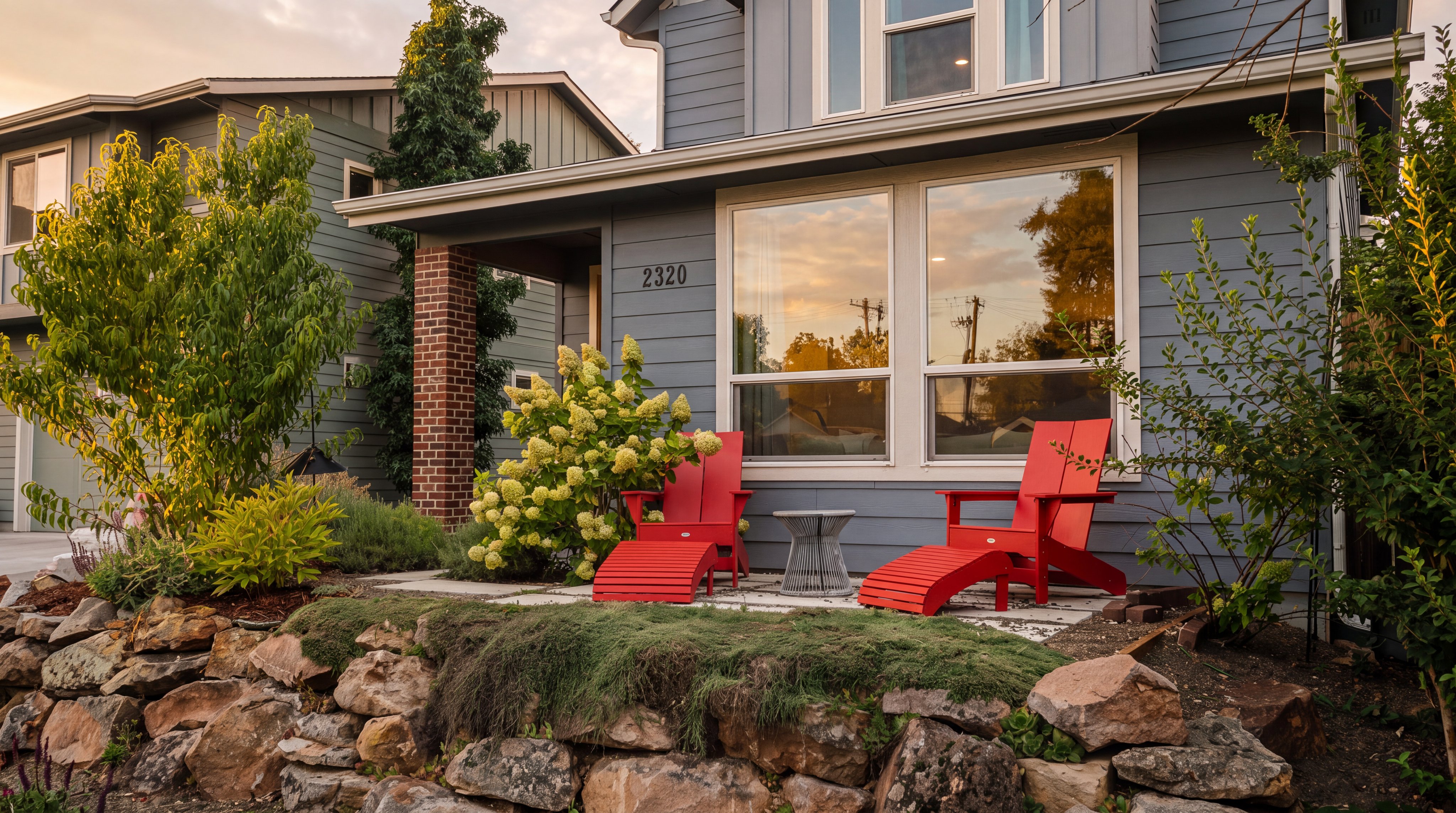 This inviting front entryway features a charming porch area with two bright red Adirondack chairs and matching footstools, set against a blue-sided home. The scene is framed by a rustic stone retaining wall in the foreground and lush, mature landscaping, including a flowering hydrangea bush. The house number '2320' is clearly displayed on the exterior wall, contributing to the home's welcoming curb appeal.