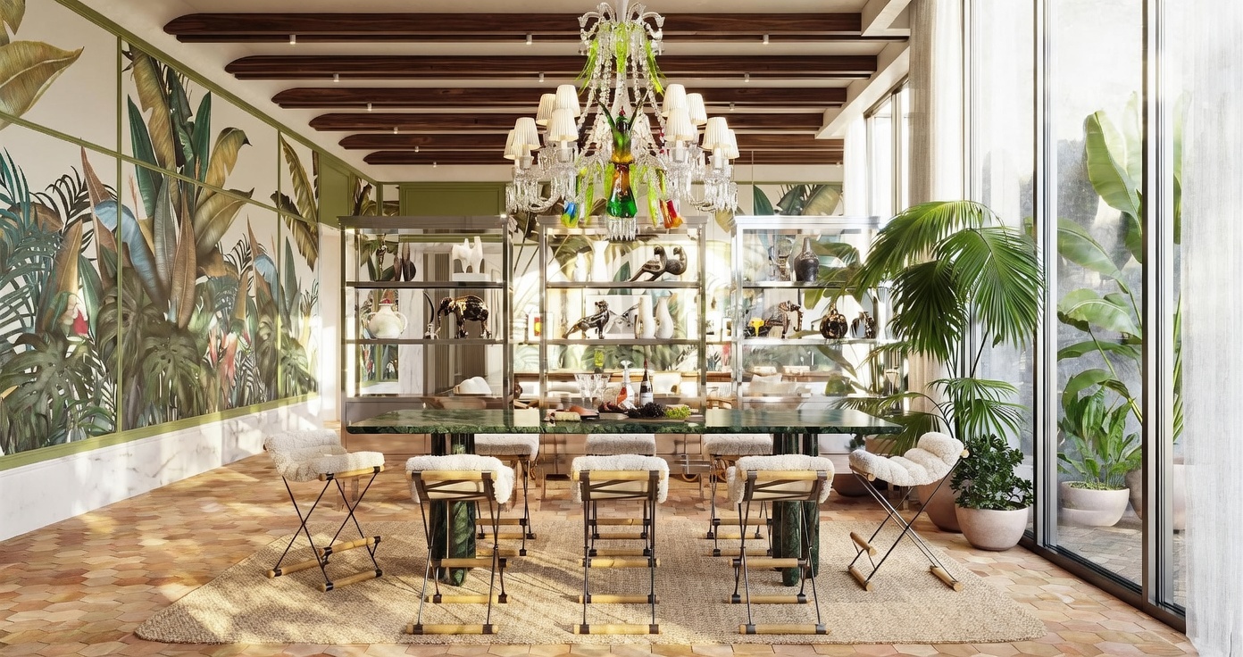This elegant dining room features a long, dark green stone table surrounded by six chairs with textured, cream-colored upholstery and metal frames. The space is anchored by a large, ornate crystal chandelier and features a striking botanical-themed wallpaper on the left wall, complemented by tall glass doors that open to an outdoor area. A central shelving unit displays decorative objects, while the room is finished with a natural fiber rug and warm, terracotta-toned floor tiles.