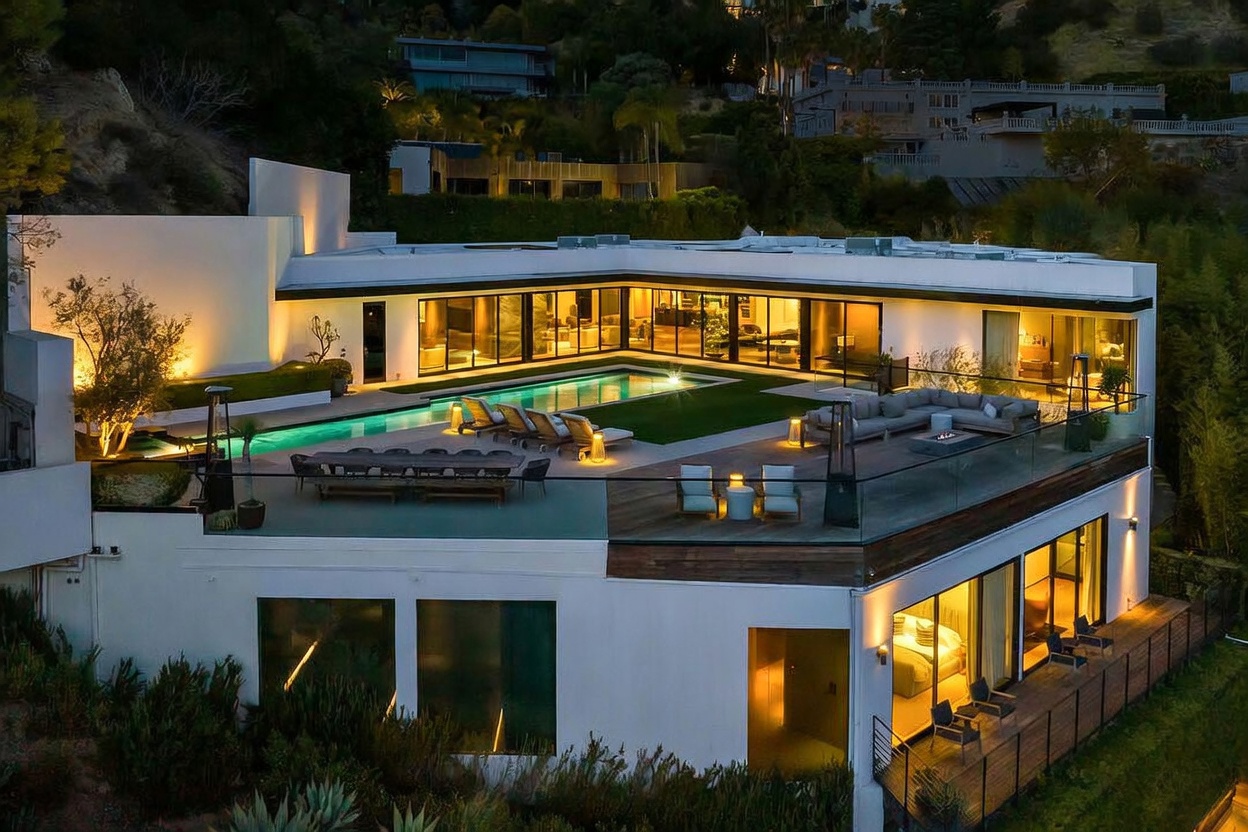 This stunning aerial view captures a modern, multi-level luxury residence at dusk, highlighting its expansive outdoor living spaces. The property features a large, illuminated swimming pool set within a manicured lawn, surrounded by a spacious deck equipped with lounge seating and an outdoor dining area. The architecture is defined by clean lines, large glass walls that provide a seamless indoor-outdoor flow, and warm ambient lighting that creates an inviting and sophisticated atmosphere.