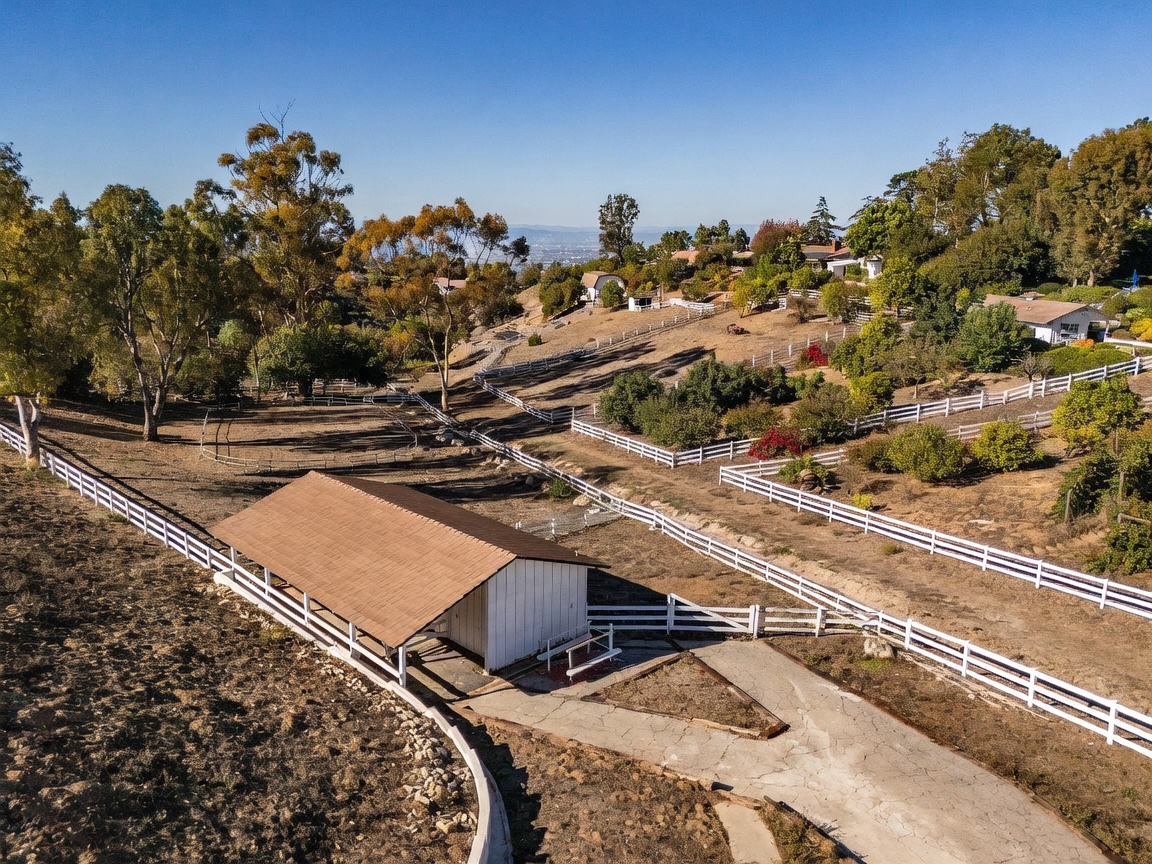 This aerial view captures a sprawling rural property featuring a white barn-style structure with a brown roof in the foreground. The landscape is characterized by rolling hills, mature trees, and extensive white fencing that delineates various sections of the land. A paved driveway leads toward the structure, providing a sense of the property's expansive and quiet setting.