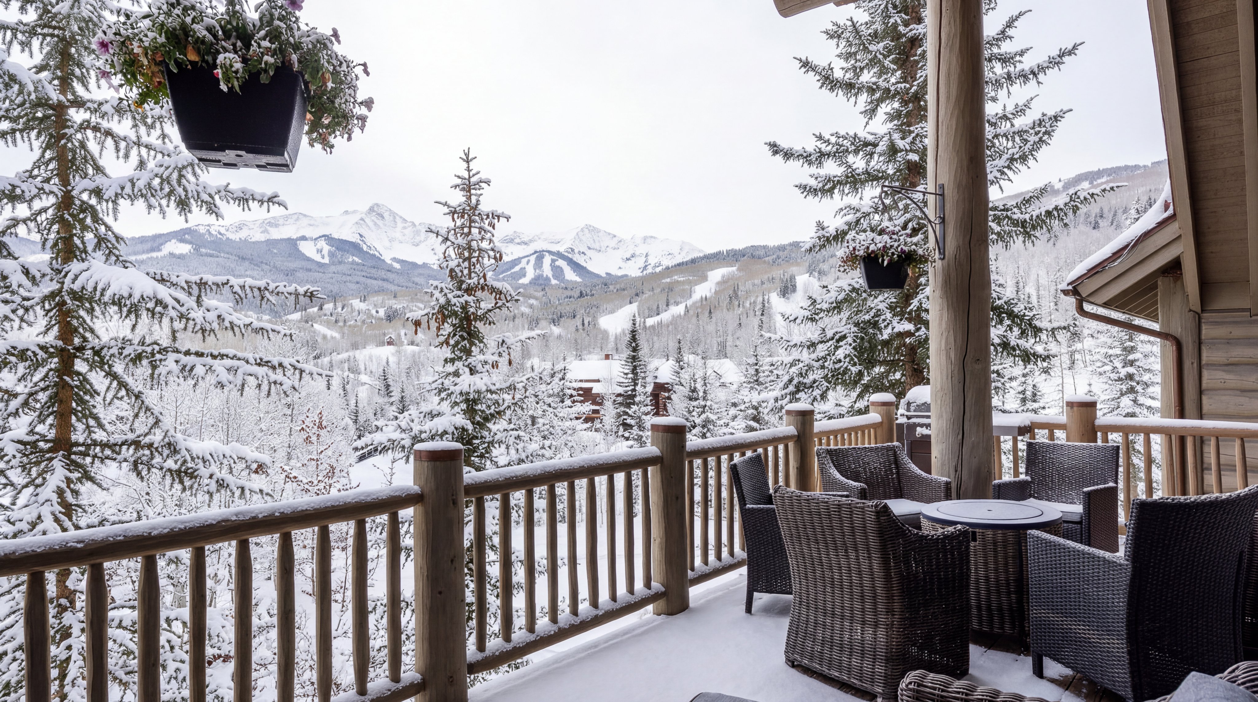 This spacious, covered balcony offers a stunning, elevated view of snow-covered mountains and a ski resort landscape. The space is furnished with comfortable, dark wicker outdoor chairs and a small table, making it an ideal spot for enjoying the serene winter scenery. The rustic wooden railing and structural pillars enhance the mountainous cabin aesthetic, creating a cozy and luxurious outdoor retreat.