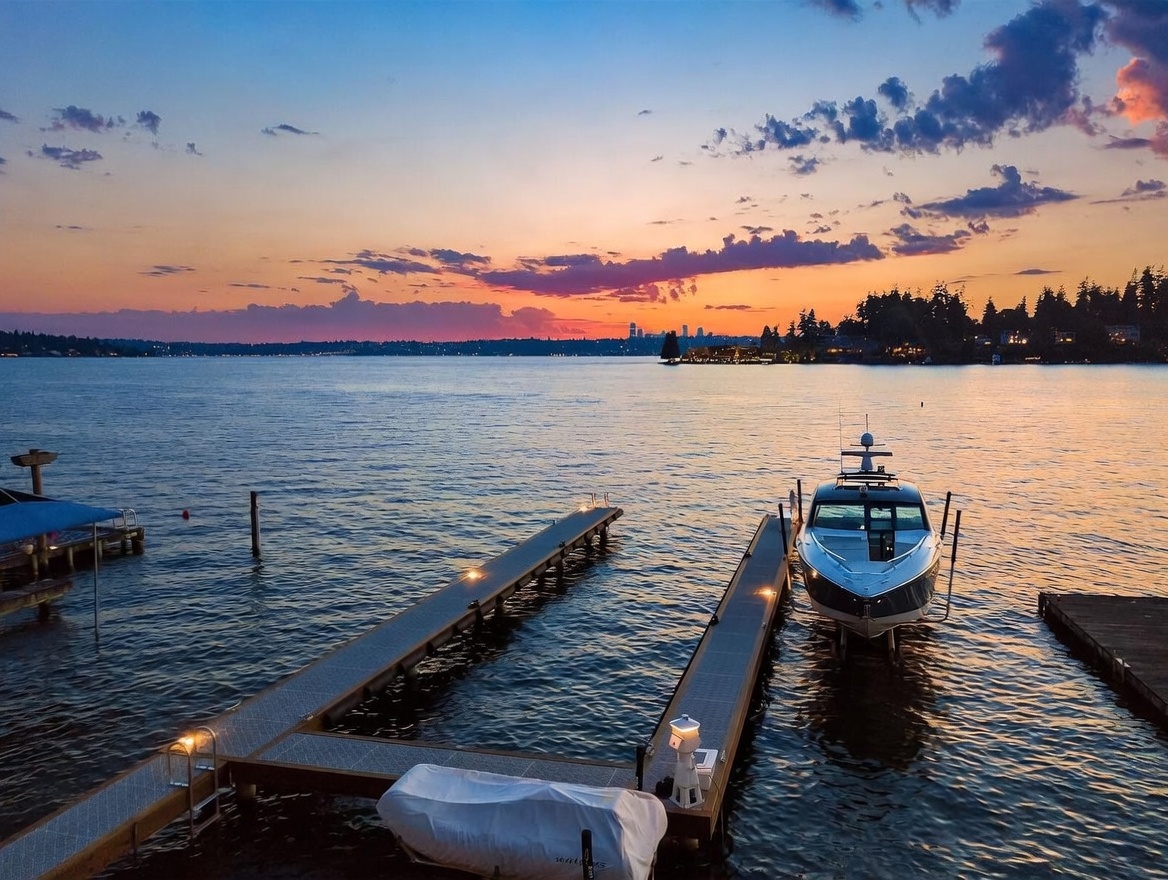 This aerial perspective captures a serene waterfront scene at sunset, featuring a private dock extending into a calm lake. A sleek motorboat is moored on one side of the dock, while a smaller, covered vessel sits at the base, all set against a vibrant, colorful sky and a distant city skyline. The composition emphasizes the tranquil lifestyle and direct water access offered by the property.