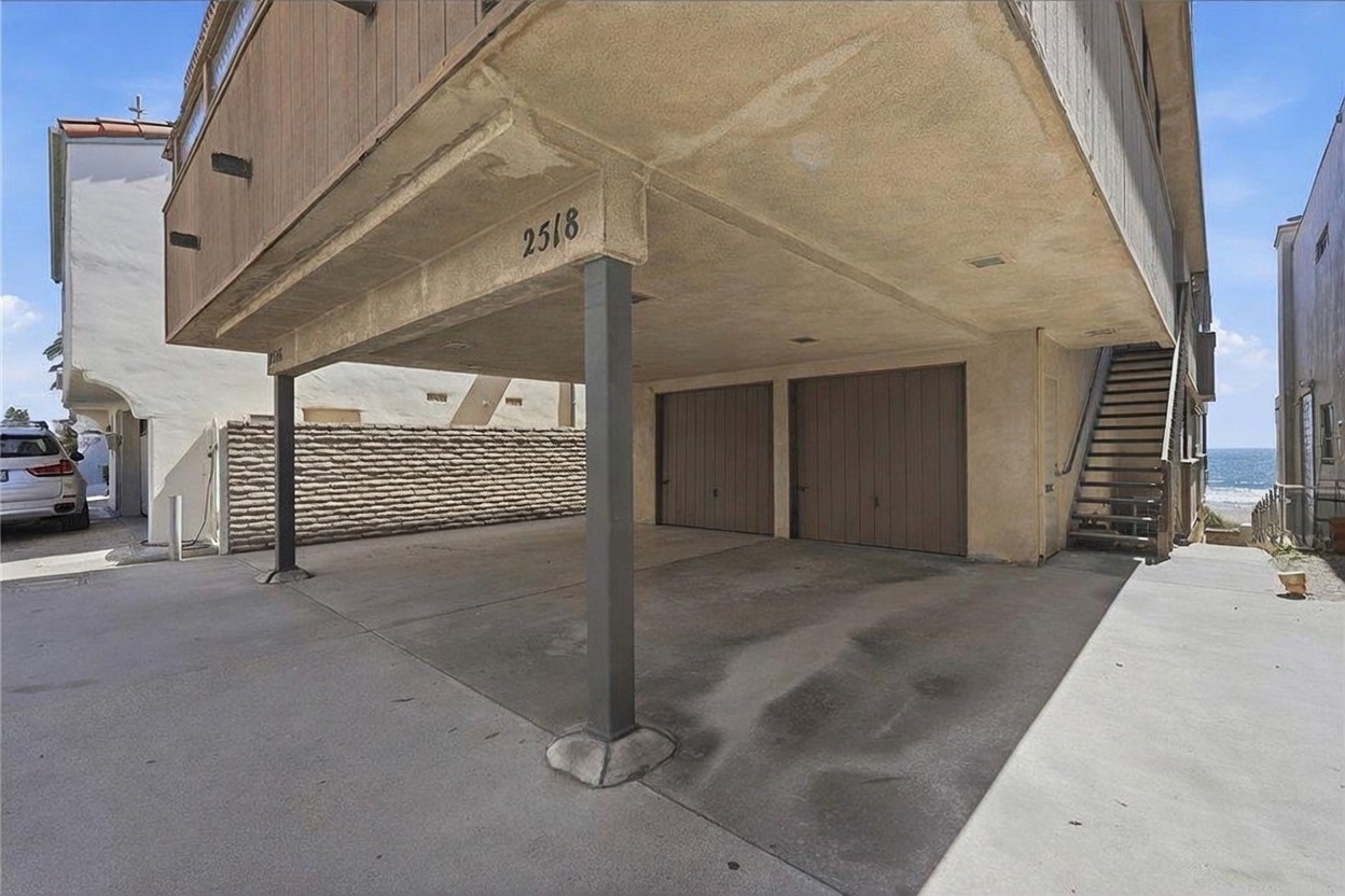 This exterior view captures the ground-level parking area of a coastal property, featuring two garage doors tucked beneath the elevated main living structure. The building is supported by sturdy vertical columns, creating an open carport space with a concrete driveway. A staircase on the right leads up to the residence, while the proximity to the ocean is visible in the background, suggesting a beachside location.