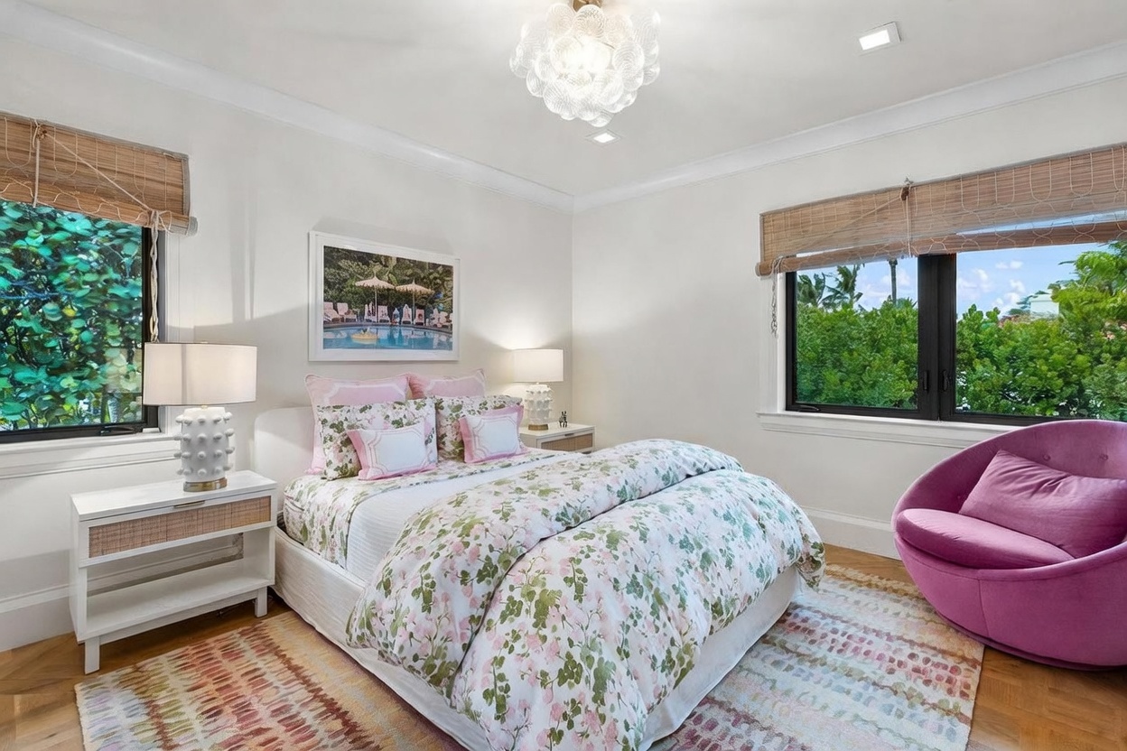 This bright and inviting bedroom features a queen-sized bed dressed in floral bedding with matching decorative pillows. A vibrant purple accent chair sits in the corner, complemented by a patterned area rug and a unique, textured white table lamp on the bedside table. Large windows with bamboo shades allow natural light to fill the room, offering a pleasant view of the lush greenery outside.