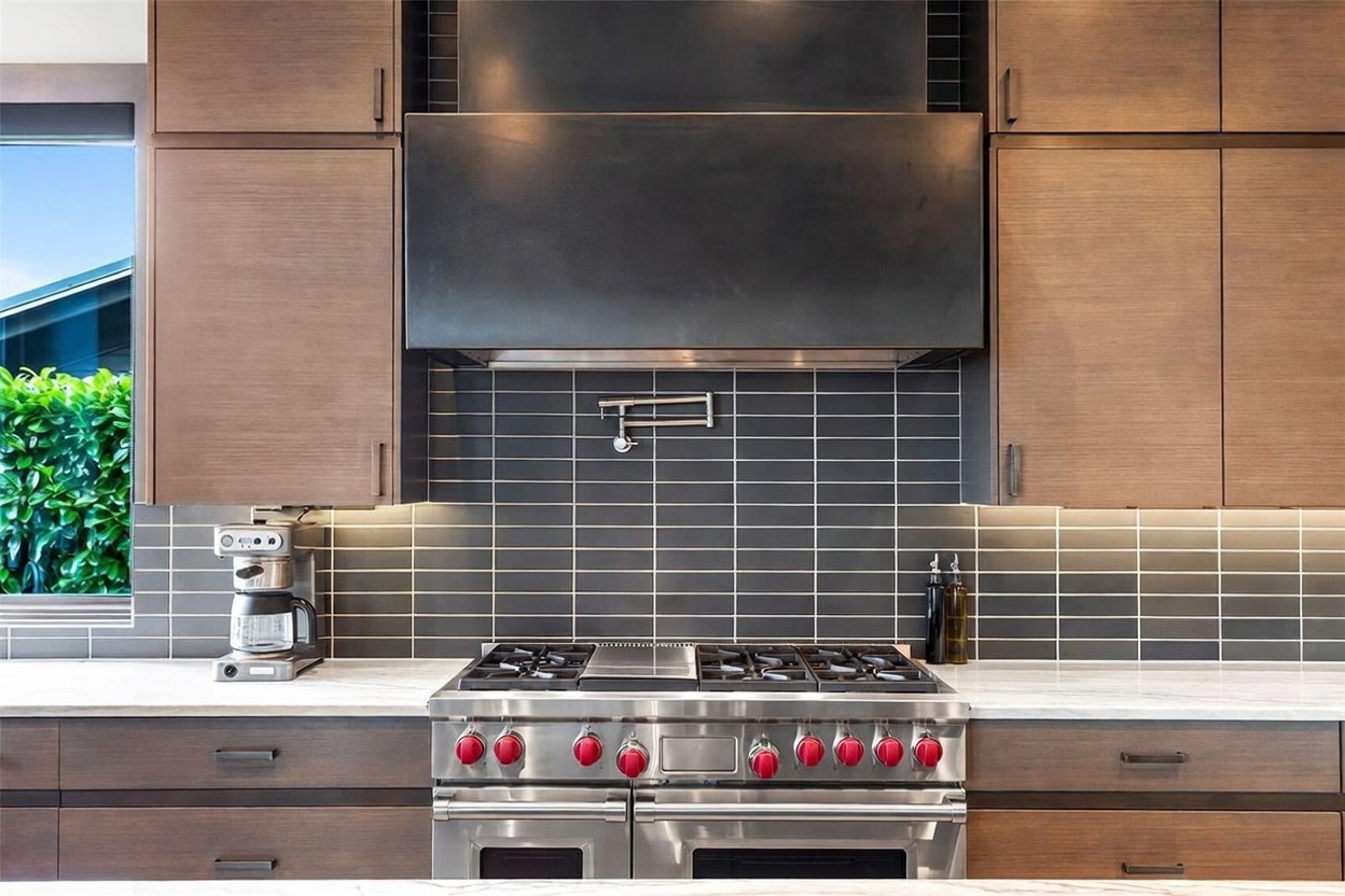 This modern kitchen features dark wood cabinetry and a professional-grade stainless steel range with a prominent hood. A dark tile backsplash provides a sleek contrast to the light-toned stone countertops, while a wall-mounted pot filler adds a functional, high-end touch. The composition is a straight-on, eye-level shot that highlights the clean lines and sophisticated, contemporary aesthetic of the culinary space.