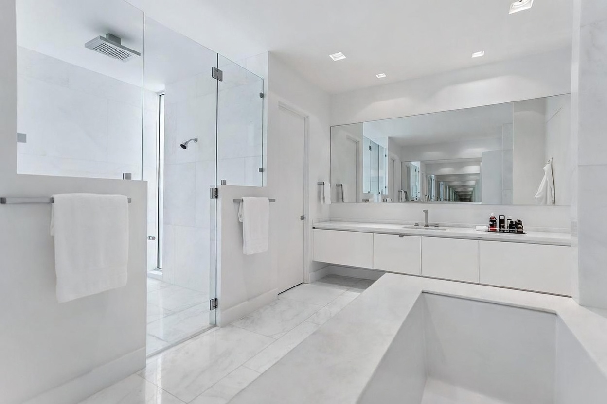 This bright, minimalist primary bathroom features a sleek, white floating vanity with a long mirror and a large, glass-enclosed walk-in shower. A deep, built-in soaking tub occupies the foreground, creating a spa-like atmosphere with its clean lines and neutral stone surfaces. The space is illuminated by recessed lighting, emphasizing the bright, airy aesthetic and high-end contemporary design.