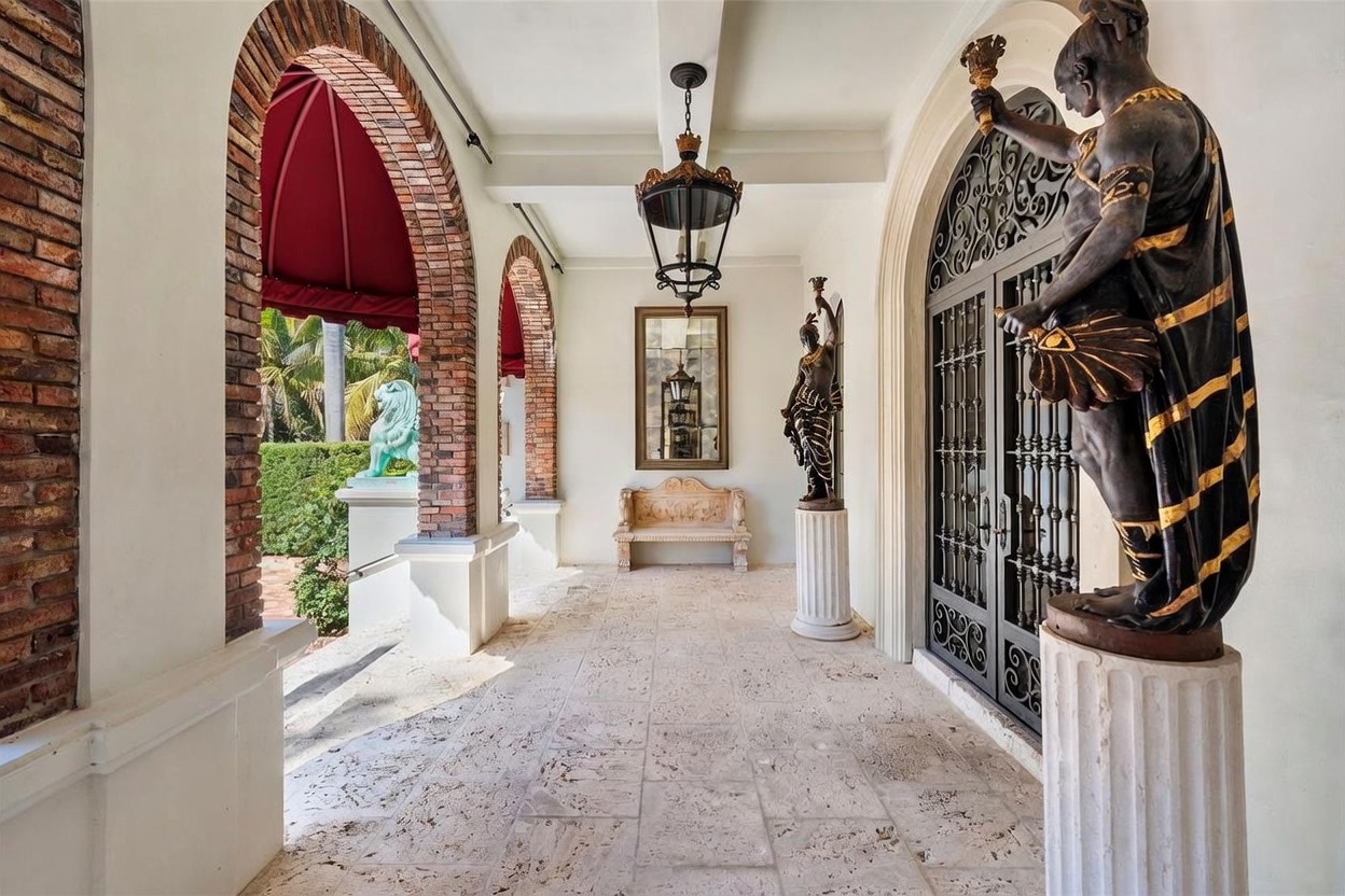 This elegant covered entryway features a stone-paved walkway framed by arched brick columns and a red fabric awning. The space is adorned with classical-style statues on pedestals, a decorative iron-grille door, and a hanging lantern, creating a sophisticated and grand atmosphere. A stone bench sits against the back wall, centered beneath a framed mirror, enhancing the formal architectural aesthetic.