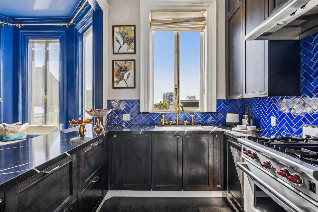 This sophisticated kitchen features dark, wood-toned cabinetry paired with striking blue herringbone-patterned tile backsplashes. A deep, dark stone countertop provides a sleek workspace, while a central window framed by white molding brings in natural light and views of the city. The space is accented by gold-toned hardware and fixtures, creating a bold and luxurious aesthetic.