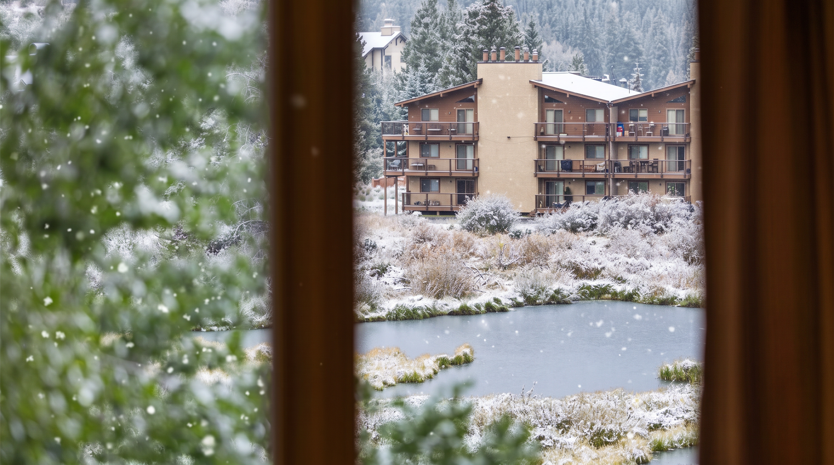 This image is captured from an interior perspective, looking through a window frame at a scenic outdoor view. The foreground is framed by blurred foliage and vertical window trim, while the background features a multi-story residential building nestled against a lush, forested hillside. A tranquil pond surrounded by tall grasses and greenery sits between the viewer and the building, creating a peaceful and picturesque setting.
