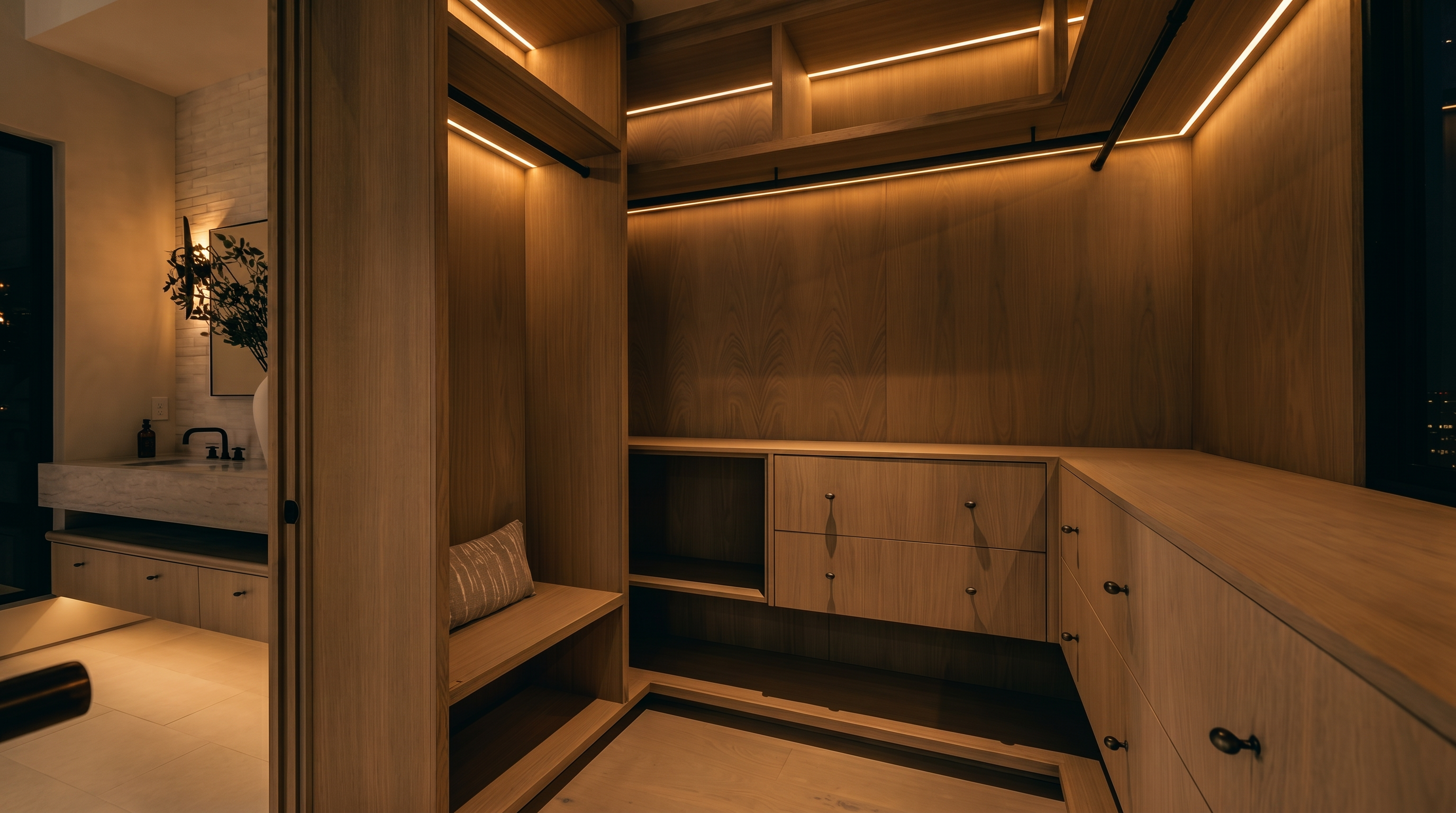This sophisticated walk-in closet features custom light wood cabinetry with integrated LED strip lighting throughout the shelving and hanging rods. The layout includes a built-in bench with a patterned cushion, multiple drawers, and open shelving, all finished in a cohesive, minimalist aesthetic. A glimpse into the adjacent bathroom reveals a stone-topped vanity and a textured wall, highlighting the seamless transition between the dressing area and the primary bath.