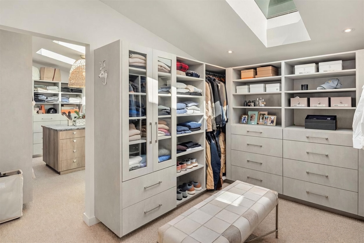 This expansive, well-organized walk-in closet features light-toned cabinetry with a mix of glass-fronted doors, open shelving, and deep drawers. A central upholstered bench provides a convenient seating area, while a secondary closet space is visible through the doorway, complete with a central island and a decorative pendant light. The room is brightened by natural light streaming through a skylight, creating a clean and luxurious atmosphere.
