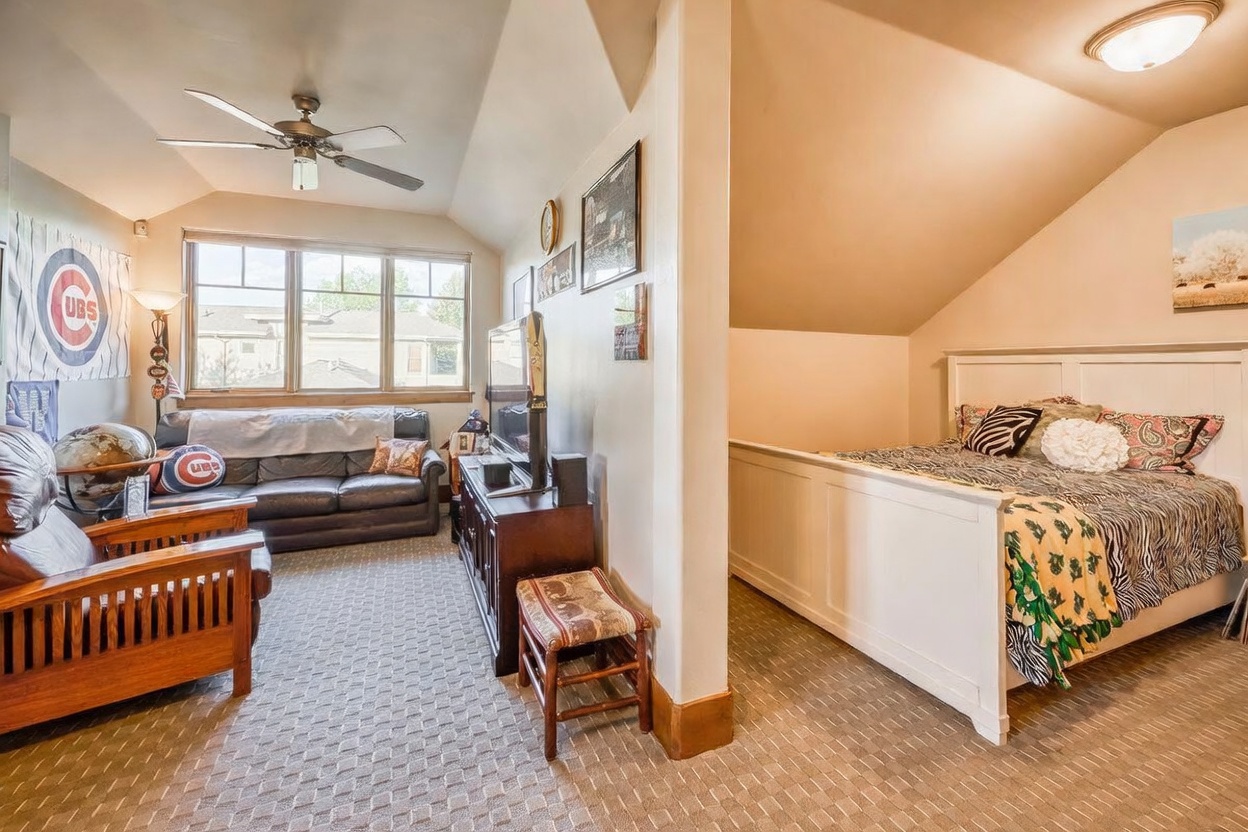 This multi-functional space features a cozy seating area with a leather sofa and a Chicago Cubs-themed decor, seamlessly transitioning into a bedroom area with a white-framed bed. The room is characterized by a vaulted ceiling, neutral carpeting, and large windows that allow for ample natural light. The layout creates a comfortable, casual living environment that serves as both a den and a sleeping quarter.