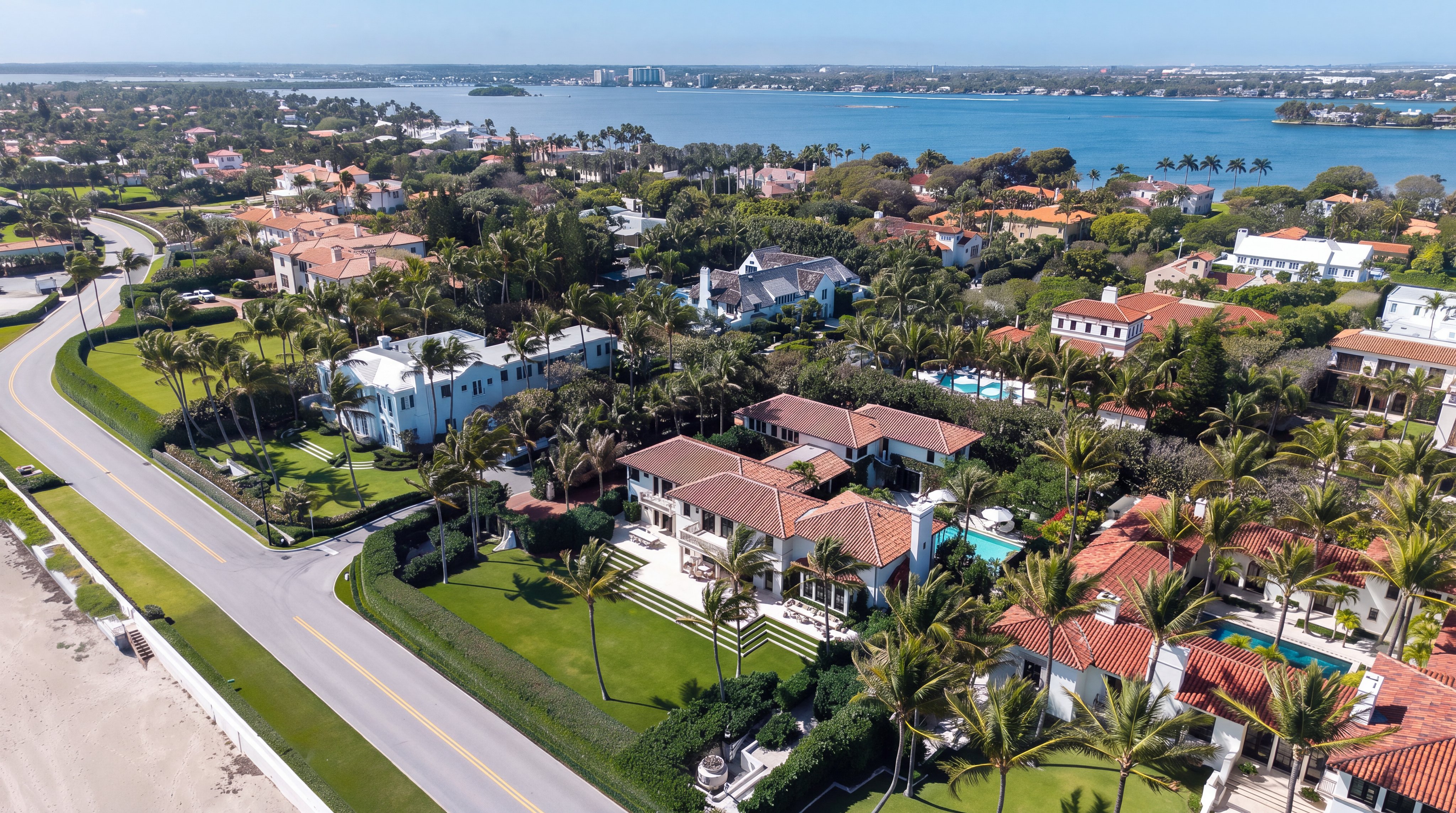 This high-angle aerial view captures a sprawling, Mediterranean-style estate situated along a coastal road with a prominent waterfront backdrop. The property features a multi-gabled tile roof, a lush green lawn, and a private swimming pool nestled within a landscaped courtyard. The perspective highlights the home's prime location, showcasing its proximity to the water and the surrounding upscale neighborhood.
