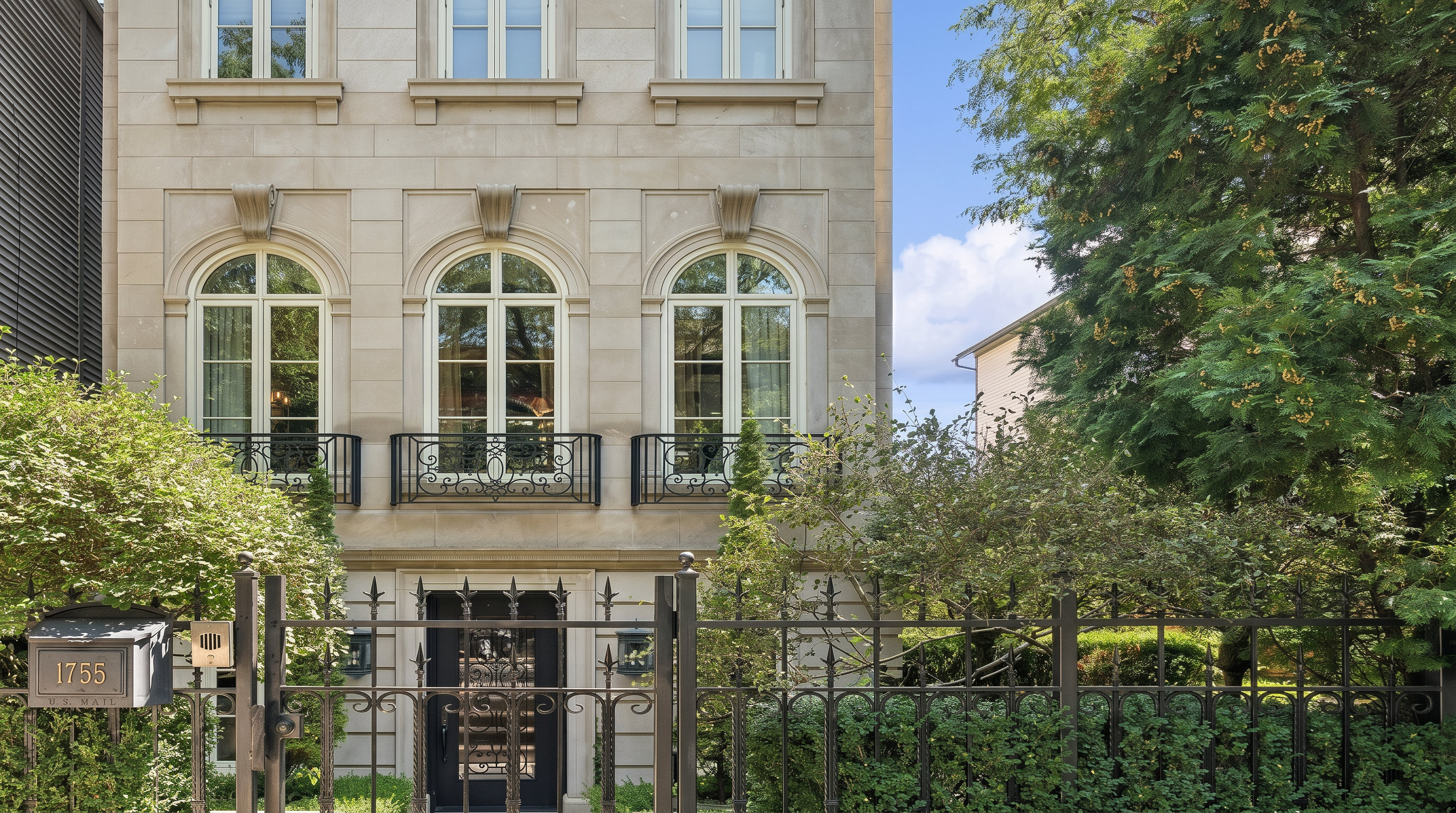 This elegant limestone-clad townhouse features a stately facade with classic arched windows and ornate black wrought-iron Juliet balconies. A decorative metal fence and a private gated entry lend an air of exclusivity and refinement to the residence. Lush surrounding greenery and a prominent mailbox marked '1755' complete this sophisticated, high-end urban aesthetic.