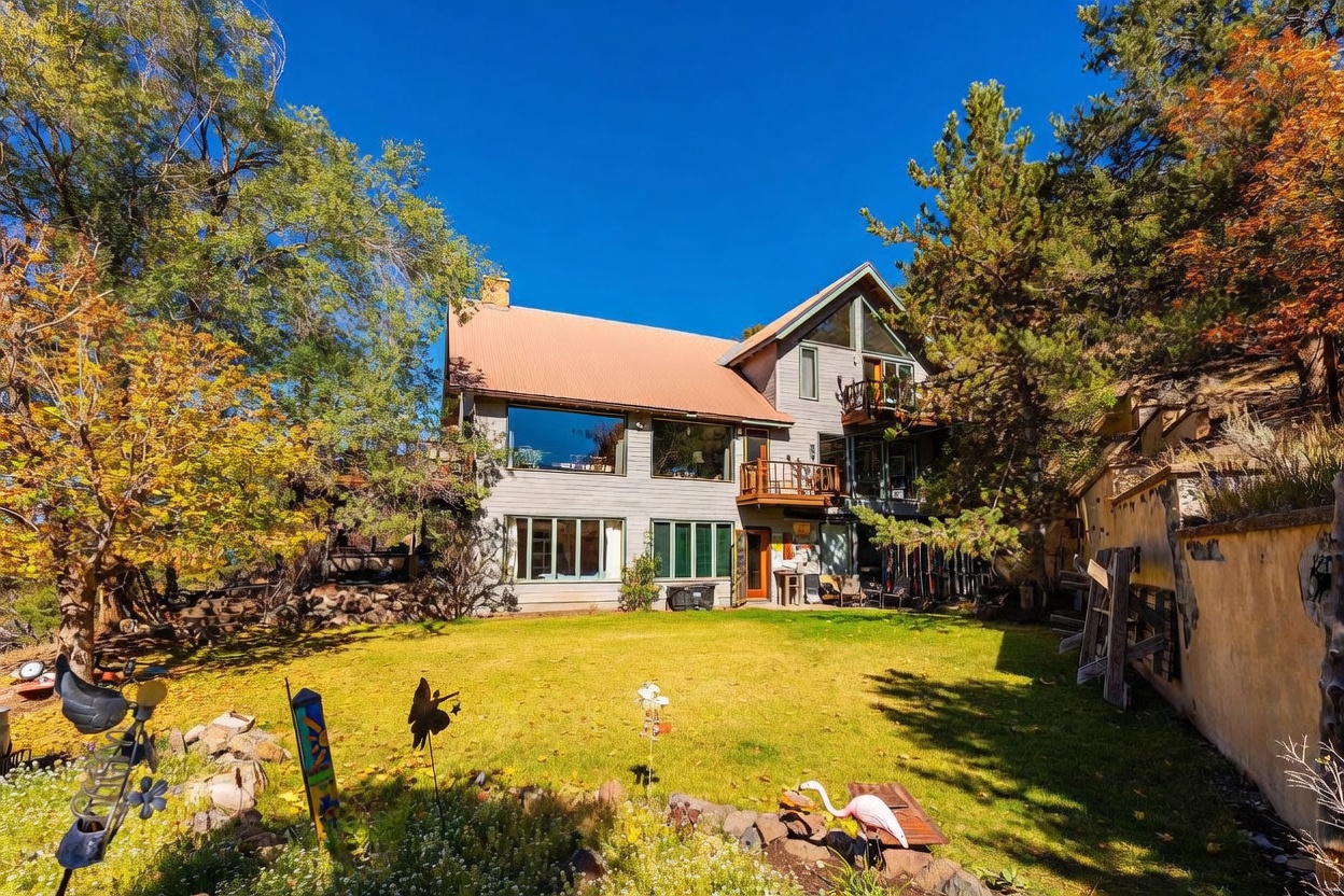 This rear view of a multi-level home showcases a rustic, mountain-inspired design with a prominent metal roof and wood-paneled siding. The house features large windows that capture natural light, multiple balconies, and a walk-out lower level that opens onto a well-maintained lawn. Surrounded by mature trees and a hillside, the property offers a serene, private outdoor setting.