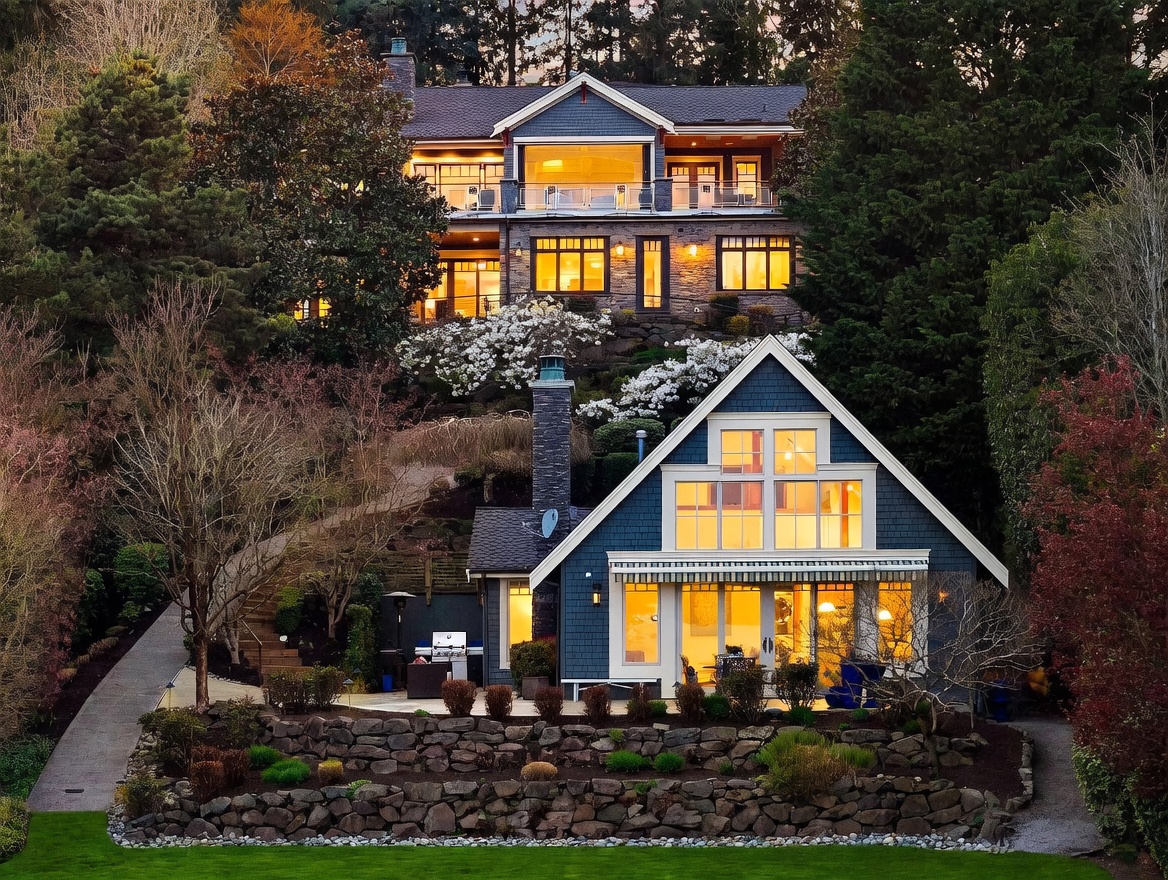 This stunning multi-level property features a primary residence perched on a hillside above a charming, cottage-style guest house or secondary living space. The homes are characterized by blue shingle siding, stone masonry accents, and expansive windows that glow warmly in the twilight. A tiered stone retaining wall and lush landscaping connect the structures, creating a picturesque and serene estate atmosphere.