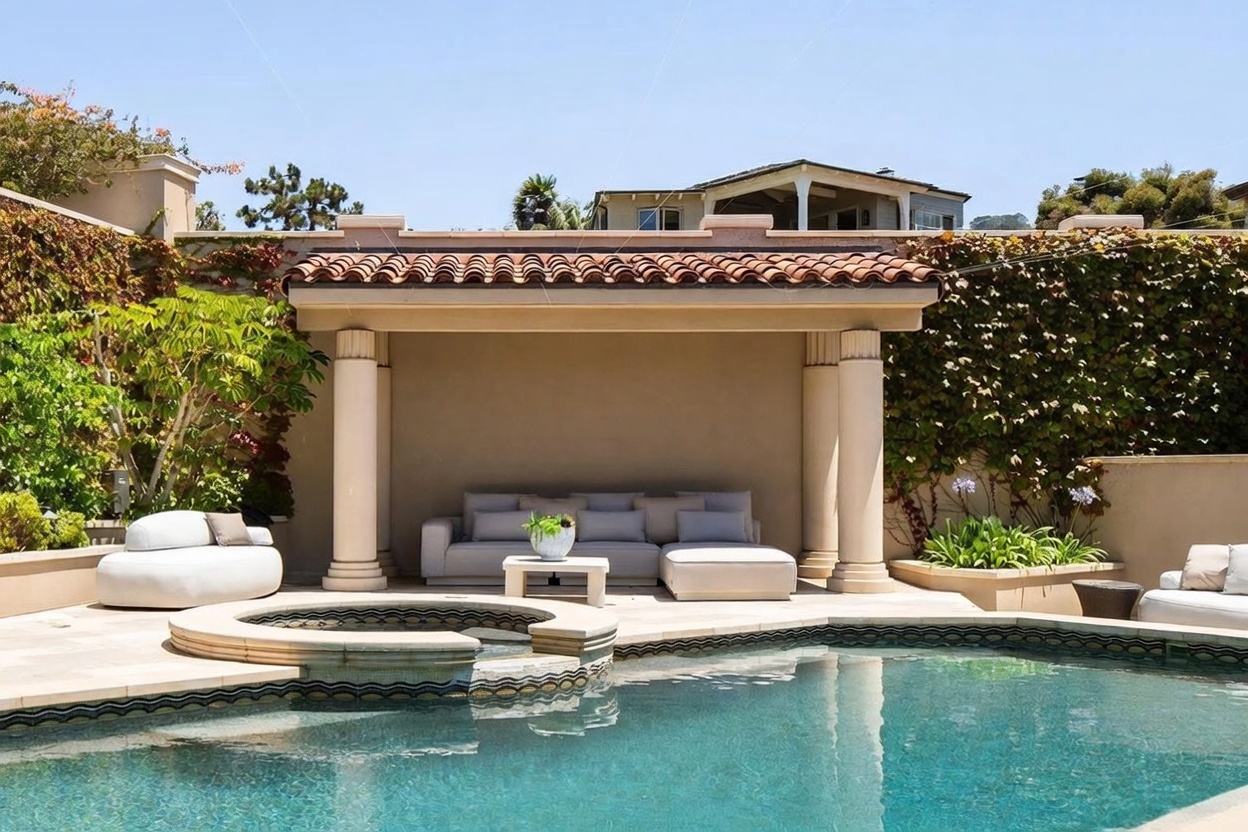 This inviting outdoor living space features a custom-shaped swimming pool with an integrated circular spa in the foreground. A covered cabana with classic columns provides a shaded lounge area furnished with a large, light-colored sectional sofa. The scene is framed by lush greenery and climbing vines on the surrounding walls, creating a private and serene resort-like atmosphere.