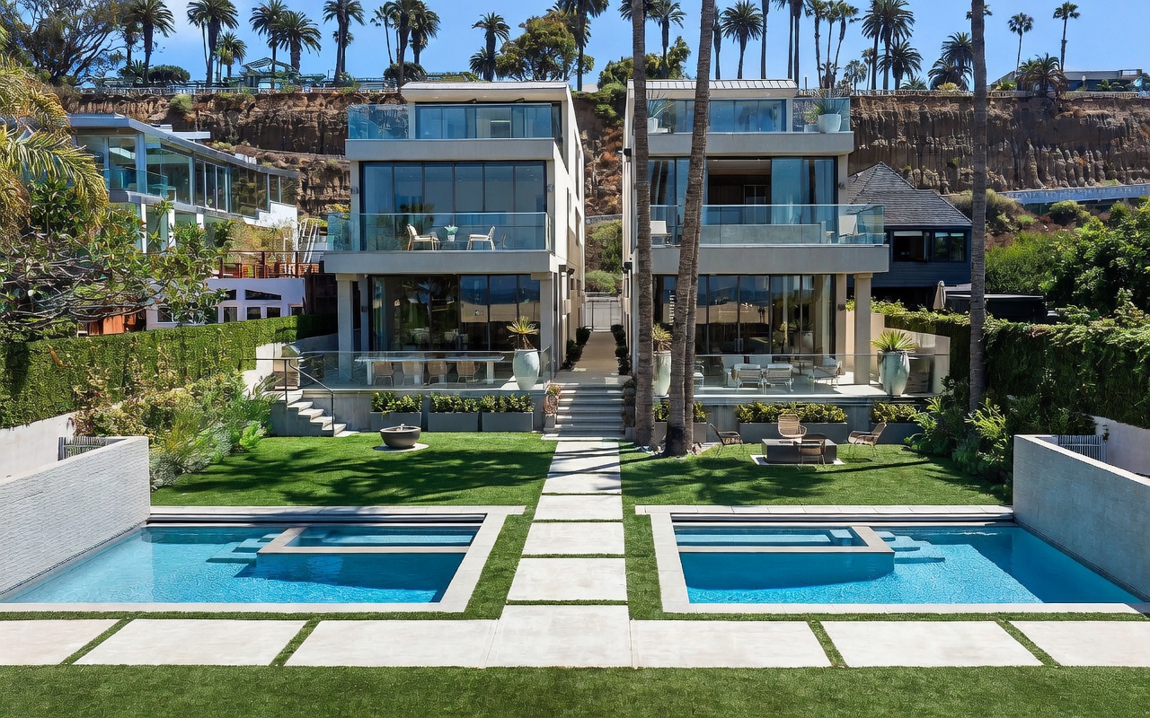 This rear view captures two modern, multi-story residences featuring expansive glass walls and tiered balconies that overlook a lush, manicured lawn. A sleek, rectangular swimming pool with integrated steps occupies the foreground, set within a clean concrete and turf patio design. The property is framed by tall palm trees and a steep, landscaped hillside, creating a private and sophisticated coastal atmosphere.