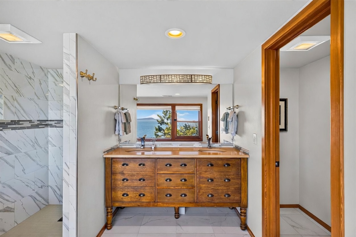 This primary bathroom features a double-sink vanity with a warm-toned wood base and a stone countertop, positioned centrally against a large mirror. To the left, a walk-in shower is enclosed by a partial wall with light-colored patterned tile, while a separate water closet is visible through a doorway on the right. The space is brightened by recessed lighting and a window above the vanity that provides a scenic view of the water.