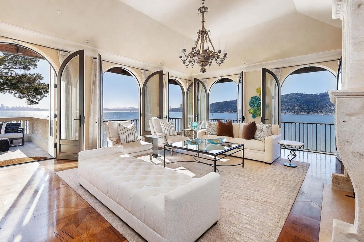 This elegant living room features a sophisticated neutral color palette, centered around a plush white tufted ottoman and a matching sofa set. The space is defined by its expansive arched glass doors that open onto a balcony, framing panoramic views of the bay and distant city skyline. A classic crystal chandelier hangs from the vaulted ceiling, adding a touch of traditional luxury to the bright, airy atmosphere.