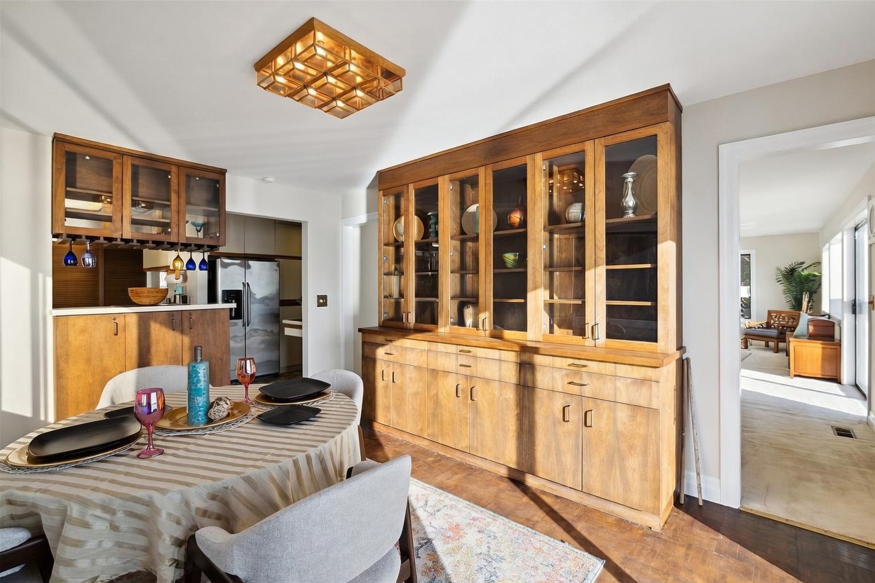 This dining room features a large, built-in wooden hutch with glass-front cabinets that provides ample storage and display space. A round dining table with a striped tablecloth sits in the foreground, surrounded by light-colored upholstered chairs. The room is illuminated by a distinctive geometric ceiling light fixture and flows into an adjacent kitchen area, creating an open and functional layout.
