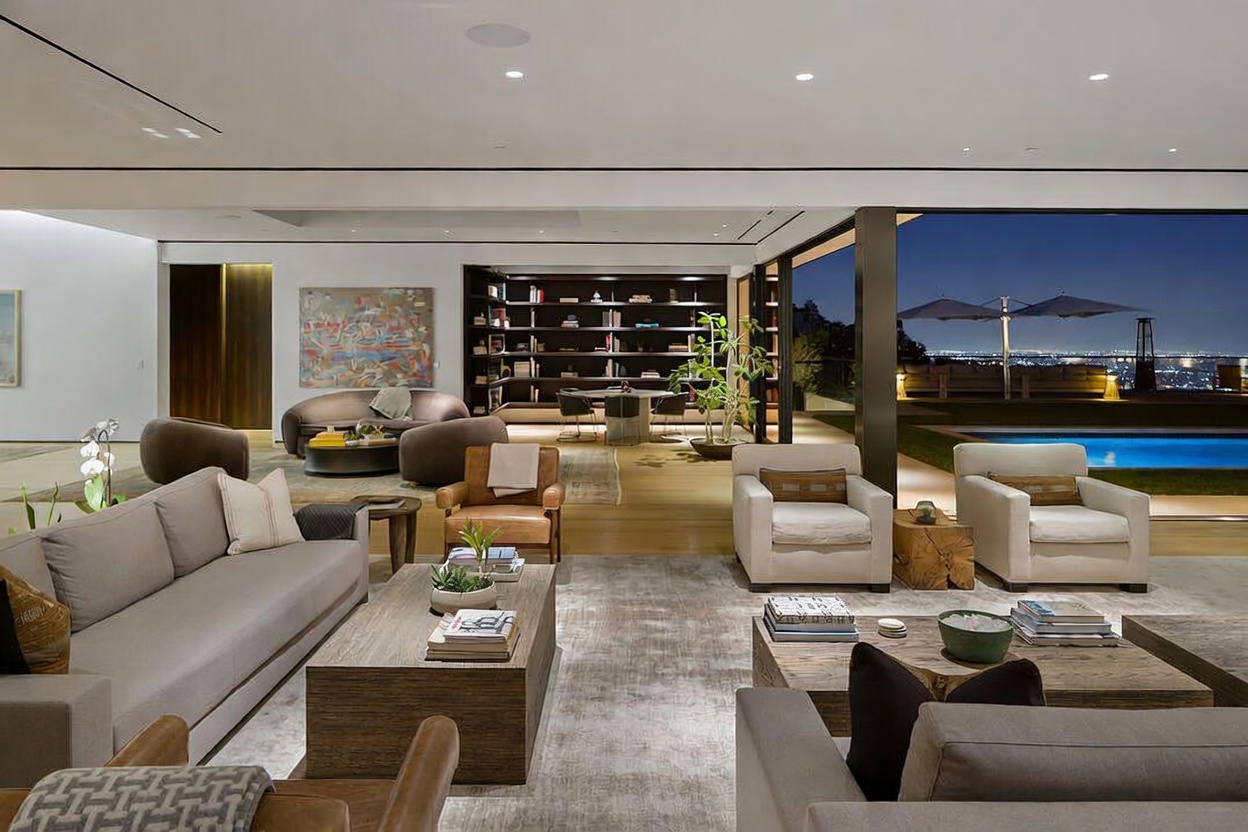This expansive, open-concept living area features a sophisticated contemporary design with neutral tones and high-end furnishings. The space seamlessly transitions to an outdoor terrace through large glass sliding doors, revealing a swimming pool and a panoramic city view at dusk. A dedicated library area with built-in shelving and a circular table sits in the background, enhancing the room's luxurious and airy atmosphere.