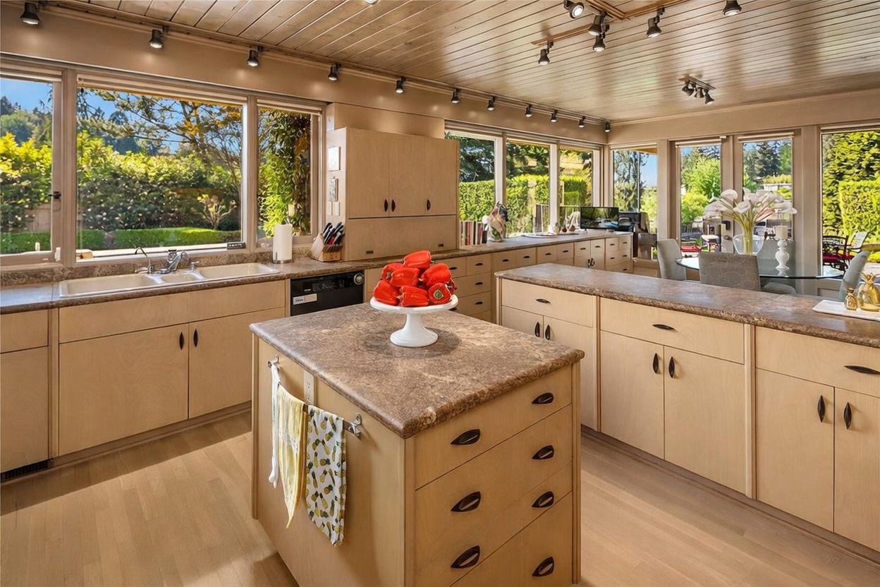 This bright and spacious kitchen features light wood cabinetry, a central island with a stone-look countertop, and a double-basin sink positioned beneath large windows that overlook a lush garden. The room is illuminated by numerous track lights mounted on a wood-paneled ceiling, creating a warm and inviting atmosphere. A dining area is visible in the background, seamlessly connecting the kitchen to the outdoor view through expansive glass panels.