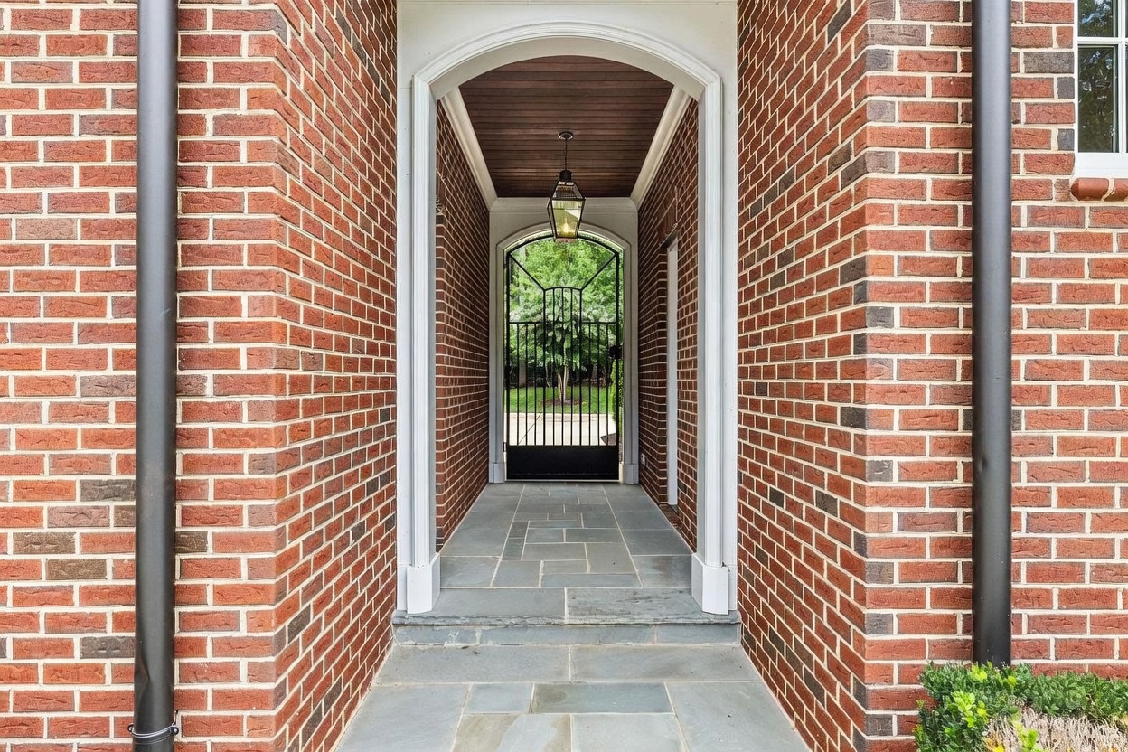 This elegant entryway features a covered walkway framed by classic red brick walls and a white arched trim. The path is paved with large, rectangular stone tiles, leading toward a decorative black metal gate that opens to a lush green outdoor space. A hanging lantern provides overhead lighting, creating a welcoming and sophisticated transition into the property.
