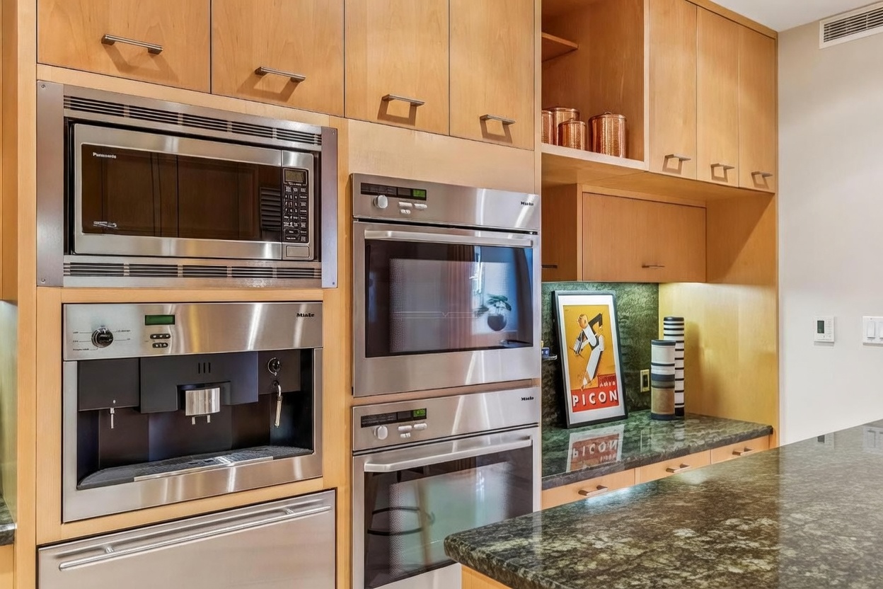 This kitchen features a wall of light wood cabinetry integrated with high-end stainless steel appliances, including a built-in coffee machine, microwave, and double ovens. To the right, a stone countertop provides a workspace or desk area, accented by a framed vintage poster and decorative canisters. The composition is captured from a straight-on perspective, highlighting the organized, professional-grade functionality of the culinary space.
