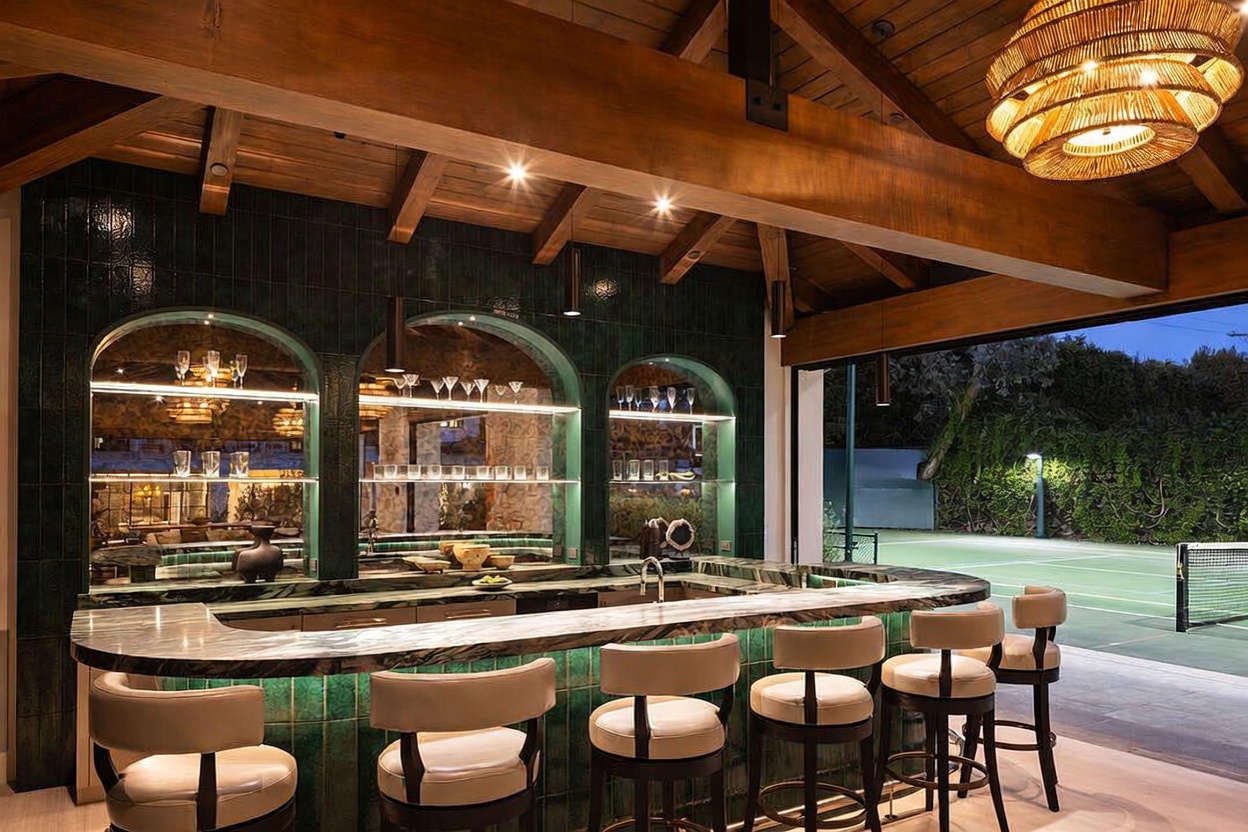 This sophisticated indoor-outdoor bar area features a curved stone-topped counter with five upholstered stools, set against a backdrop of deep green tiled walls with arched, illuminated shelving. The space is defined by exposed wooden ceiling beams and a large, textured pendant light, while the open-air design provides a seamless transition to an adjacent tennis court. The perspective captures the bar from a slightly elevated angle, emphasizing the luxurious, club-like atmosphere of this entertainment space.